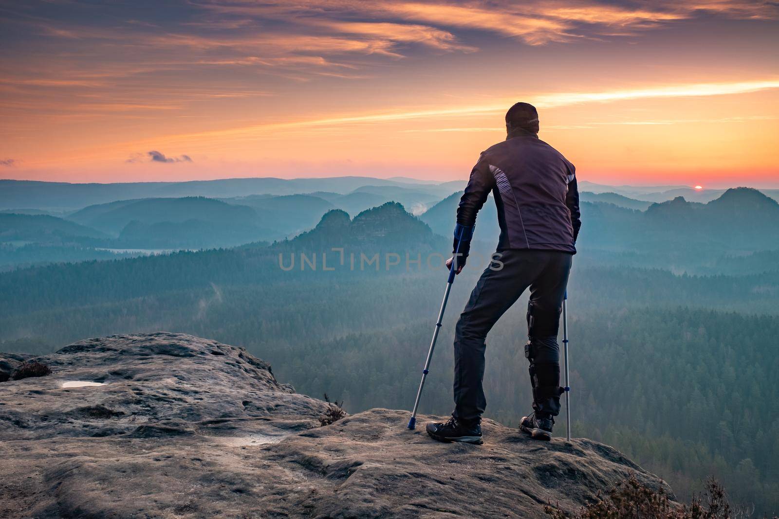 Disabled tourist with crutches. Rear view of Hurt hiker man with forearm poles walking against mountain sunset background 