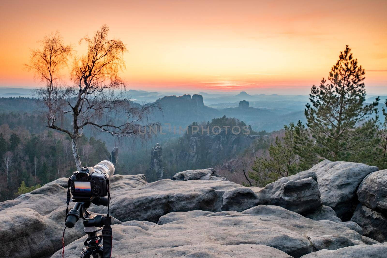 Camera on tripod is taking the sun sets just behind the Schrammsteine rock massif, Saxon Switzerland, Germany. View from the Carolafelsen viewpoint.