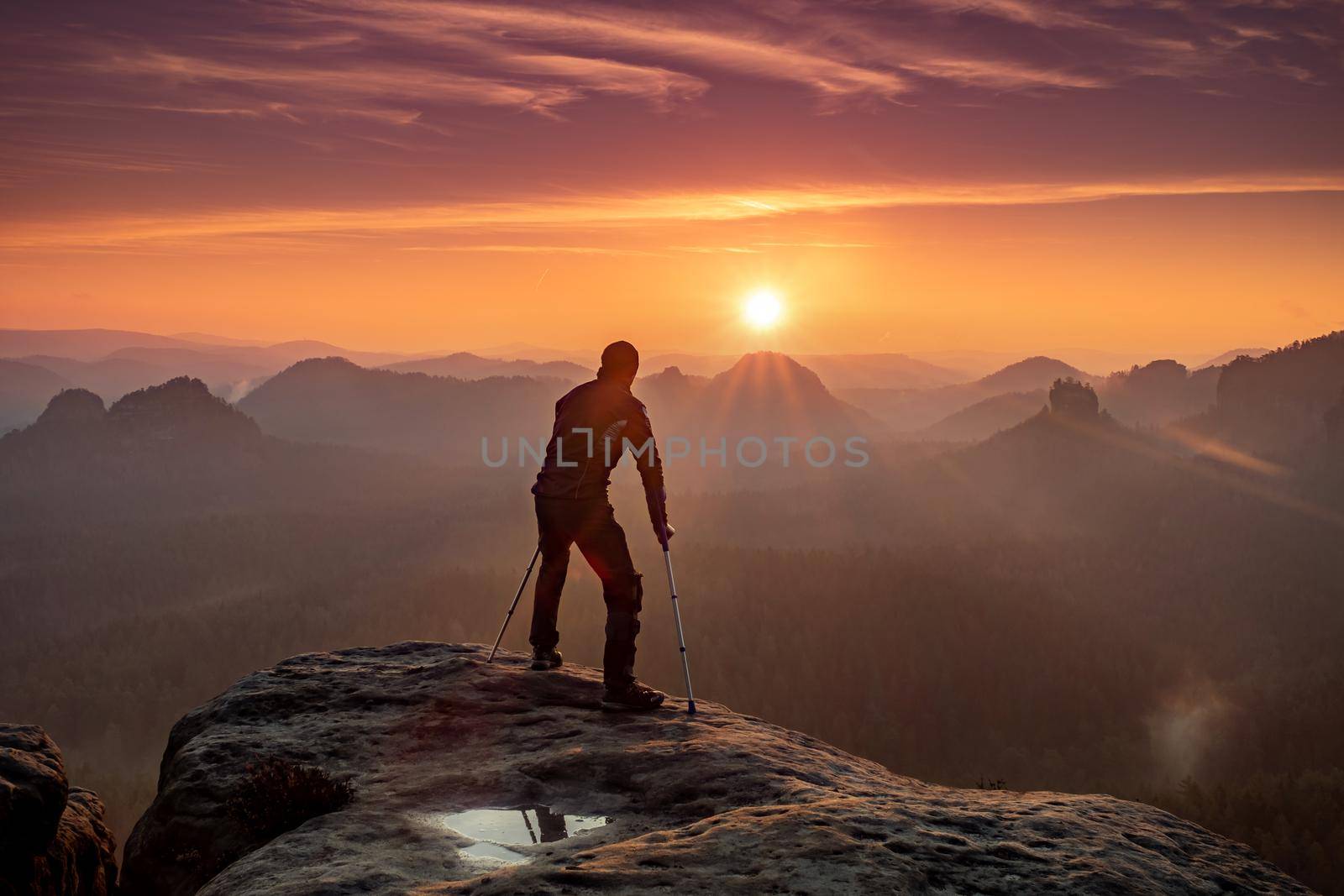 Disabled tourist with crutches. Rear view of Hurt hiker man with forearm poles walking against mountain sunset background 