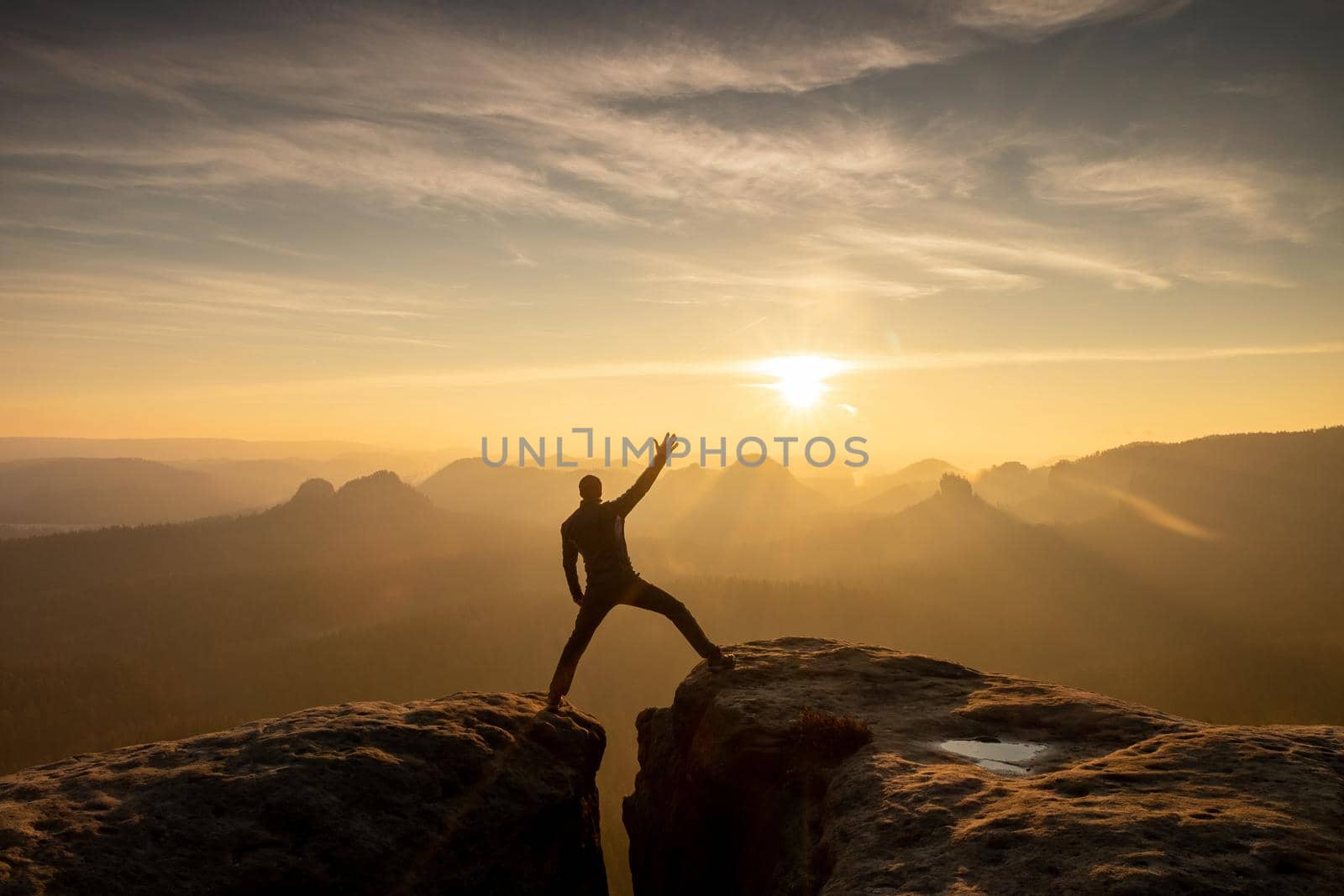 Young man standing on a cliff edge celebrates reaching the top of the mountain, arms in the air, shouting.