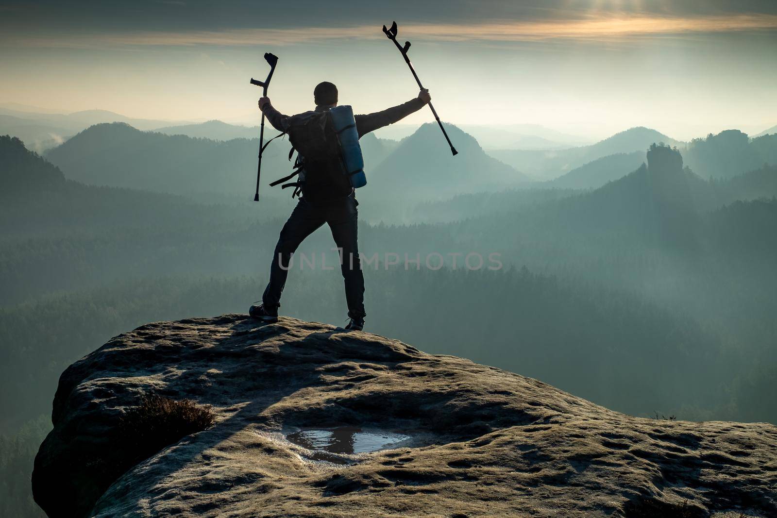 Silhouette of happy man with arms outstretched holding crutches standing by mountain valley  during sunset. Personal motivation