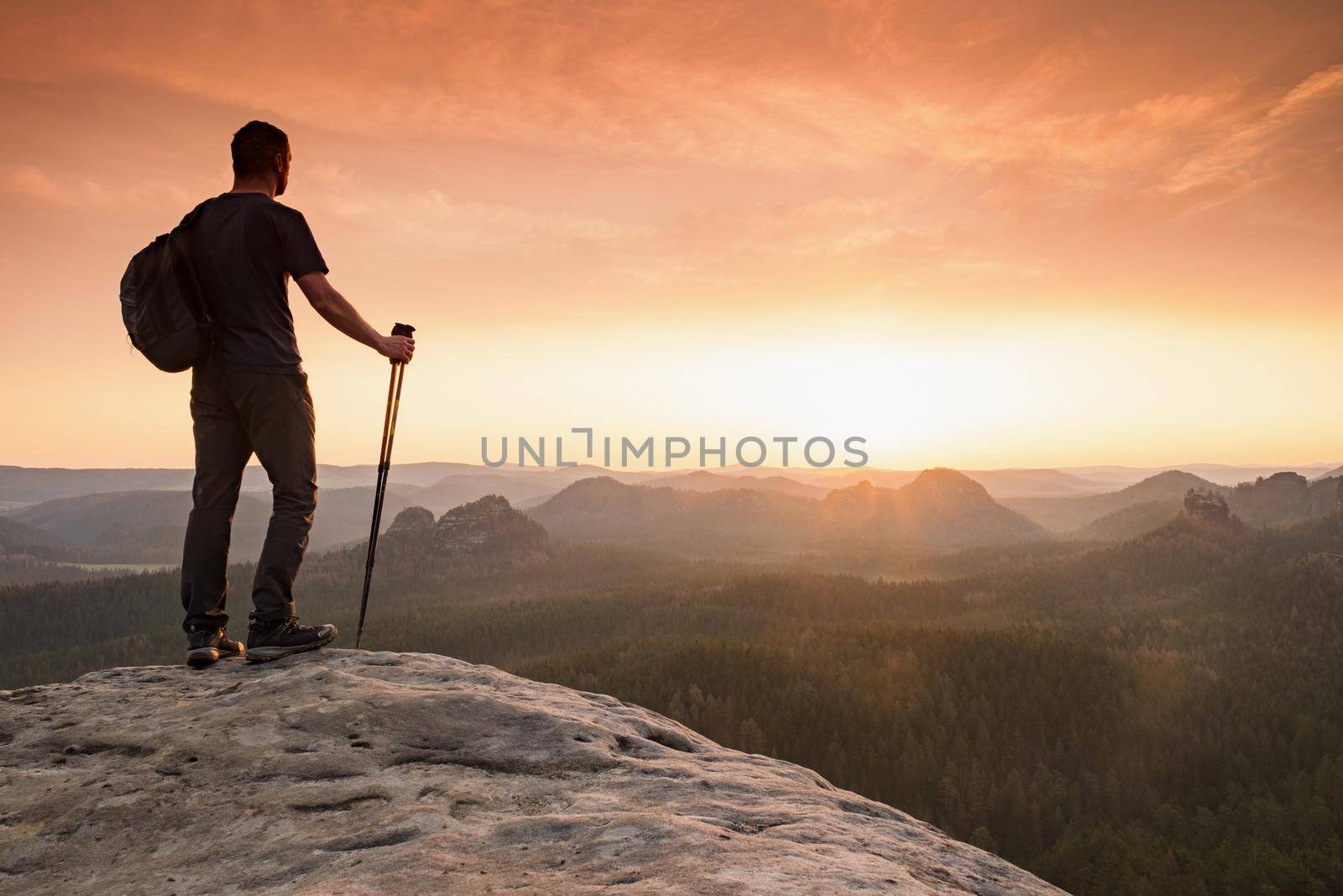 Hiker with backpack leaning on trekking poles staring into misty sunrise of Saechsische Schweiz park. Beautiful morning valley full of thick fog in orange colors. Saxony Switzerland, landscape Germany