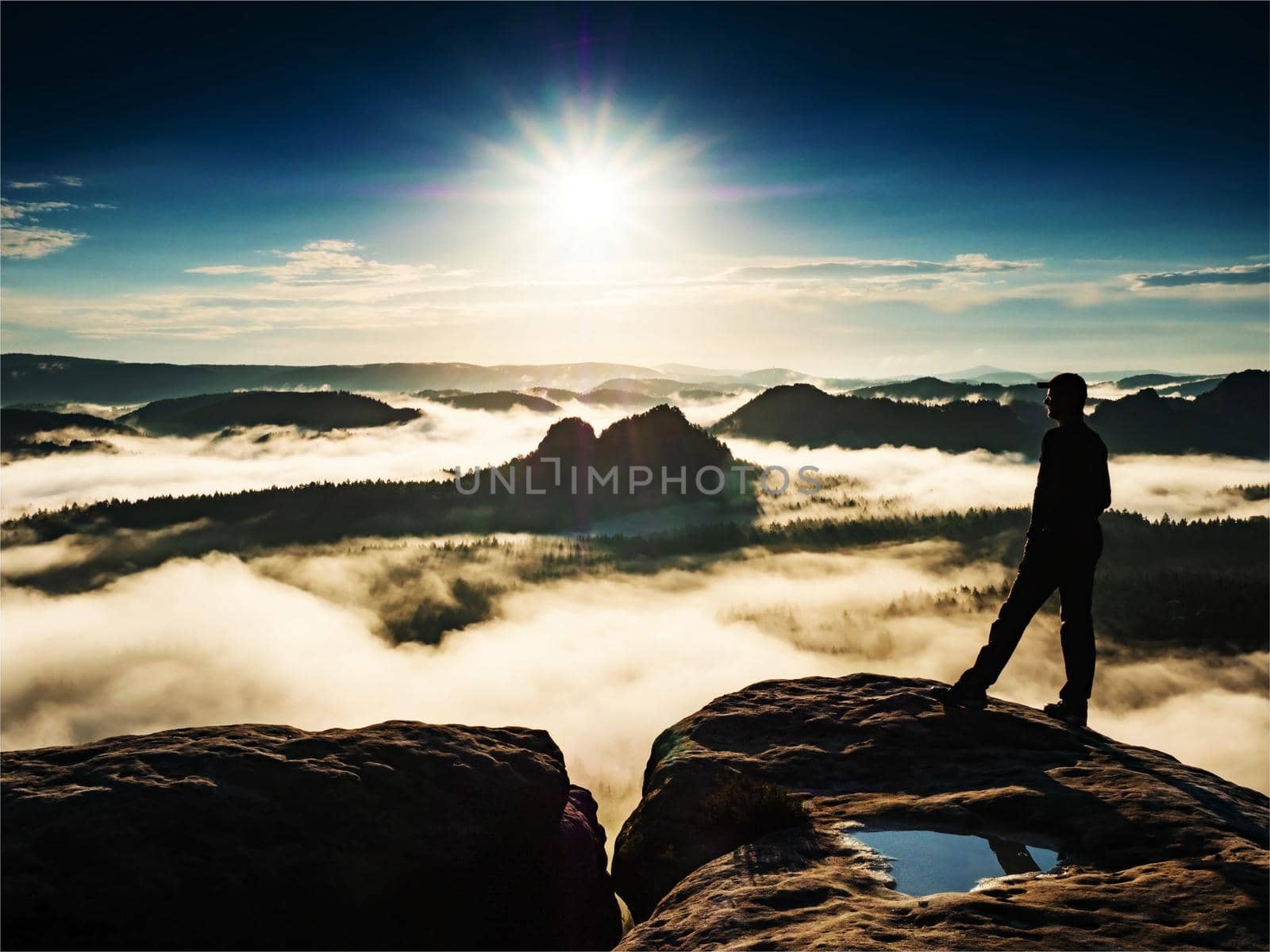 Man standing on a rock sorrounded by clouds on trek summit. Winterberg rock in the Saxon Switzerland National Park and photographs the sunrise.