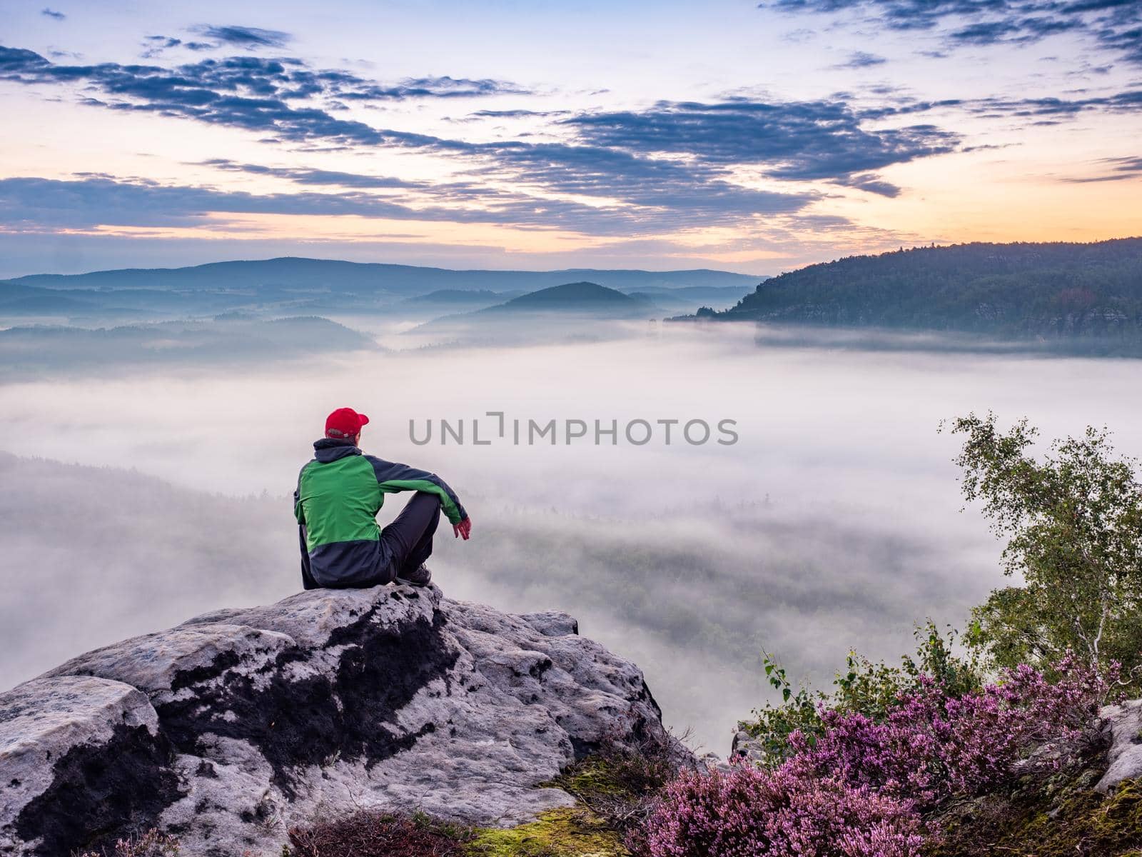 Tired hiker sits on the edge of the cliff and looking at the sun valley. Adventure un misty mountains