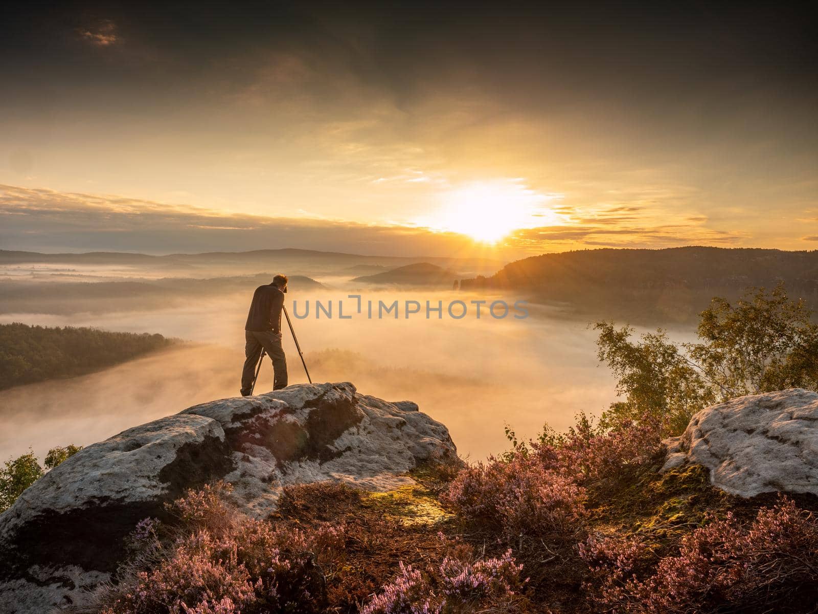 Landscape photographer in action taking picture. Silhouette man photographing against misty landscape and morning sky. Autumn colorful sunset