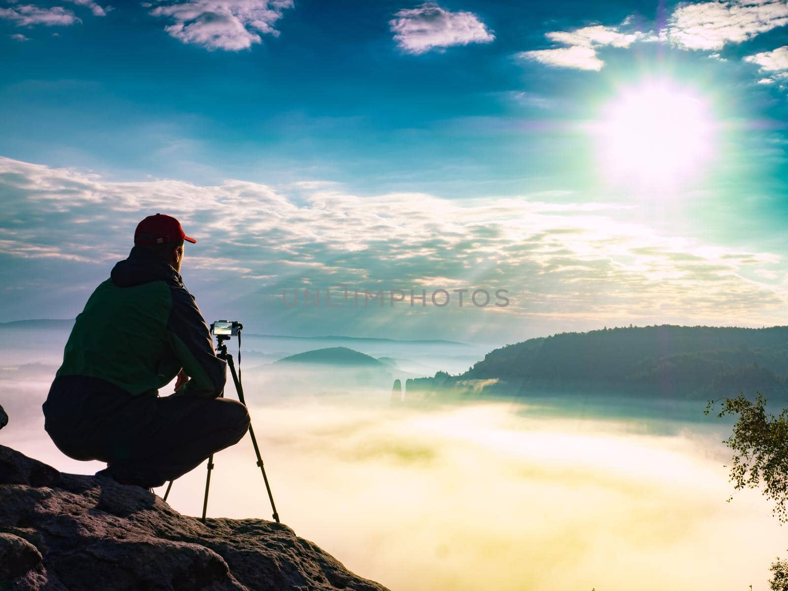 Matured man photographer at the hilltop with his tripod and camera search of good object to make a final photography session at sunset time.