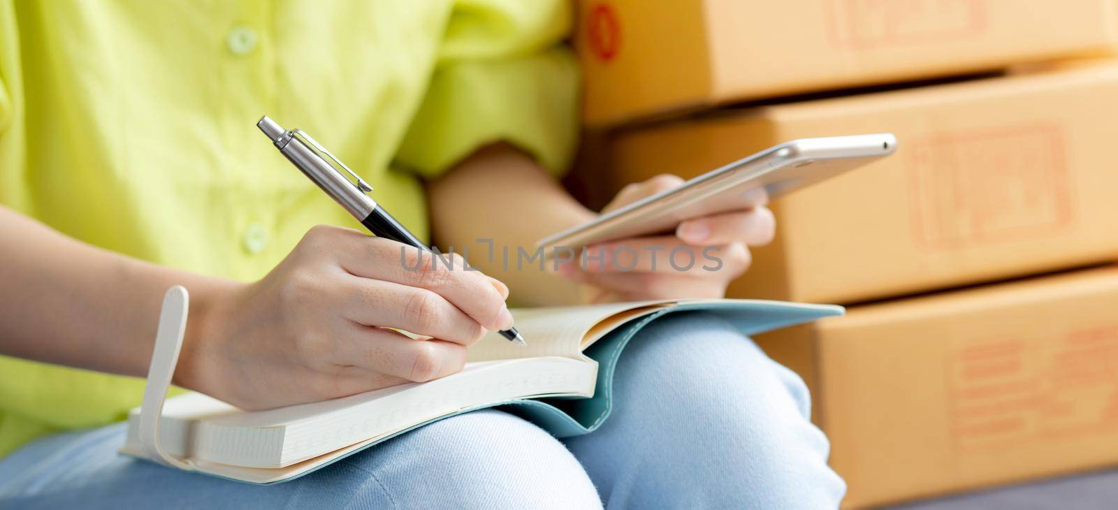 Closeup young asian woman is entrepreneur looking phone and writing order of customer with online on notebook, online shopping, logistic and delivery, female is seller working on sofa, business SME.