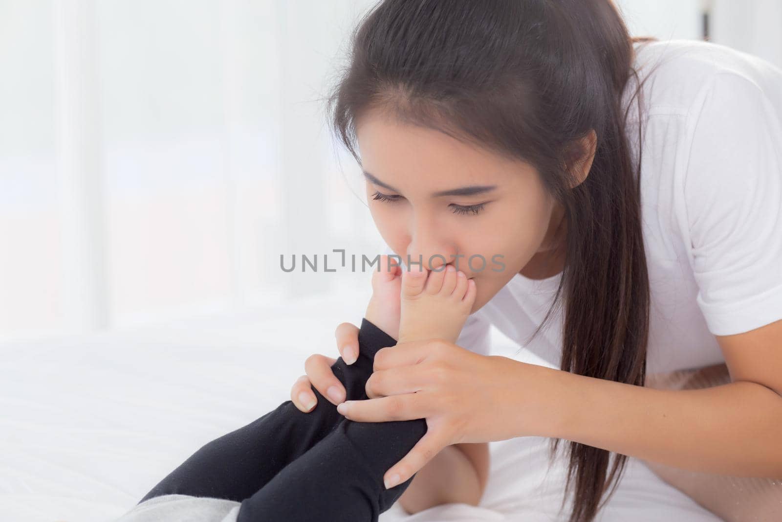 Young asian mother kiss foot of little baby girl with tender on bed in the bedroom, mom love newborn and care, mother with expression with child together, parent and daughter, family concept.