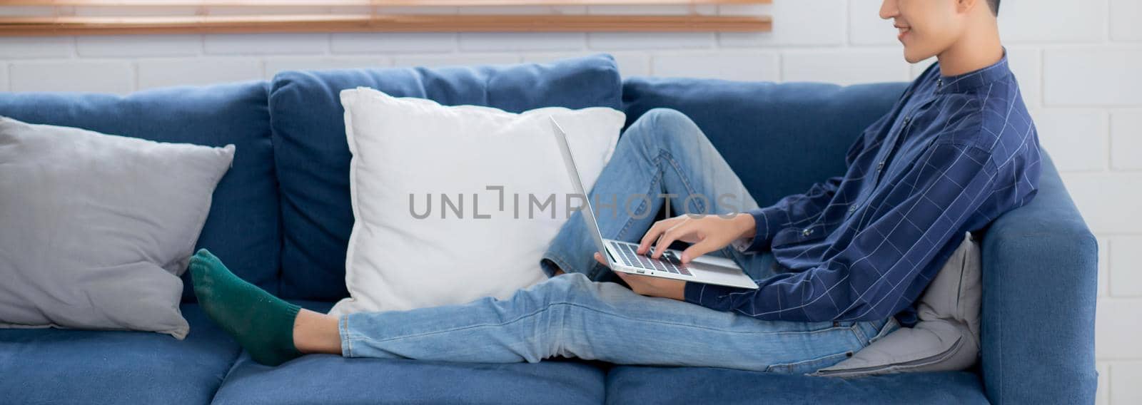 Young asian businessman smile and work from home with laptop computer online to internet on sofa in living room, freelance man using notebook on couch with comfort, new normal, lifestyle concept.