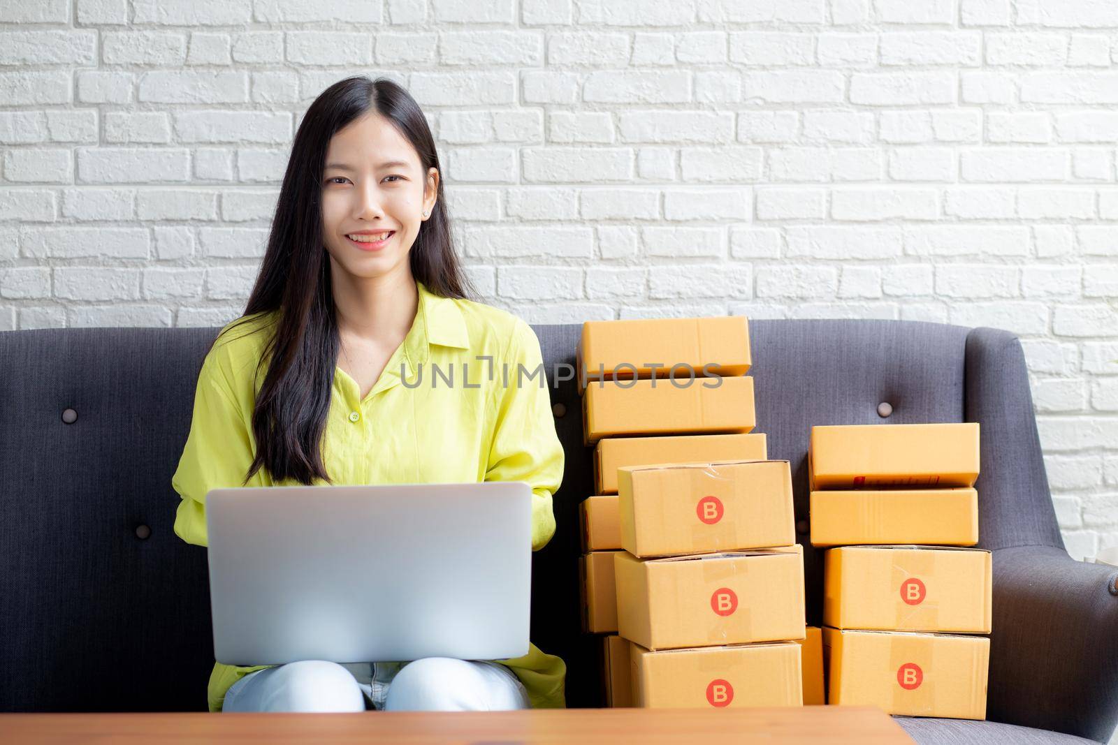 Young asian woman is merchant using laptop computer on sofa checking order of customer with purchase online shopping, business SME or startup, entrepreneur or seller and service delivery product.