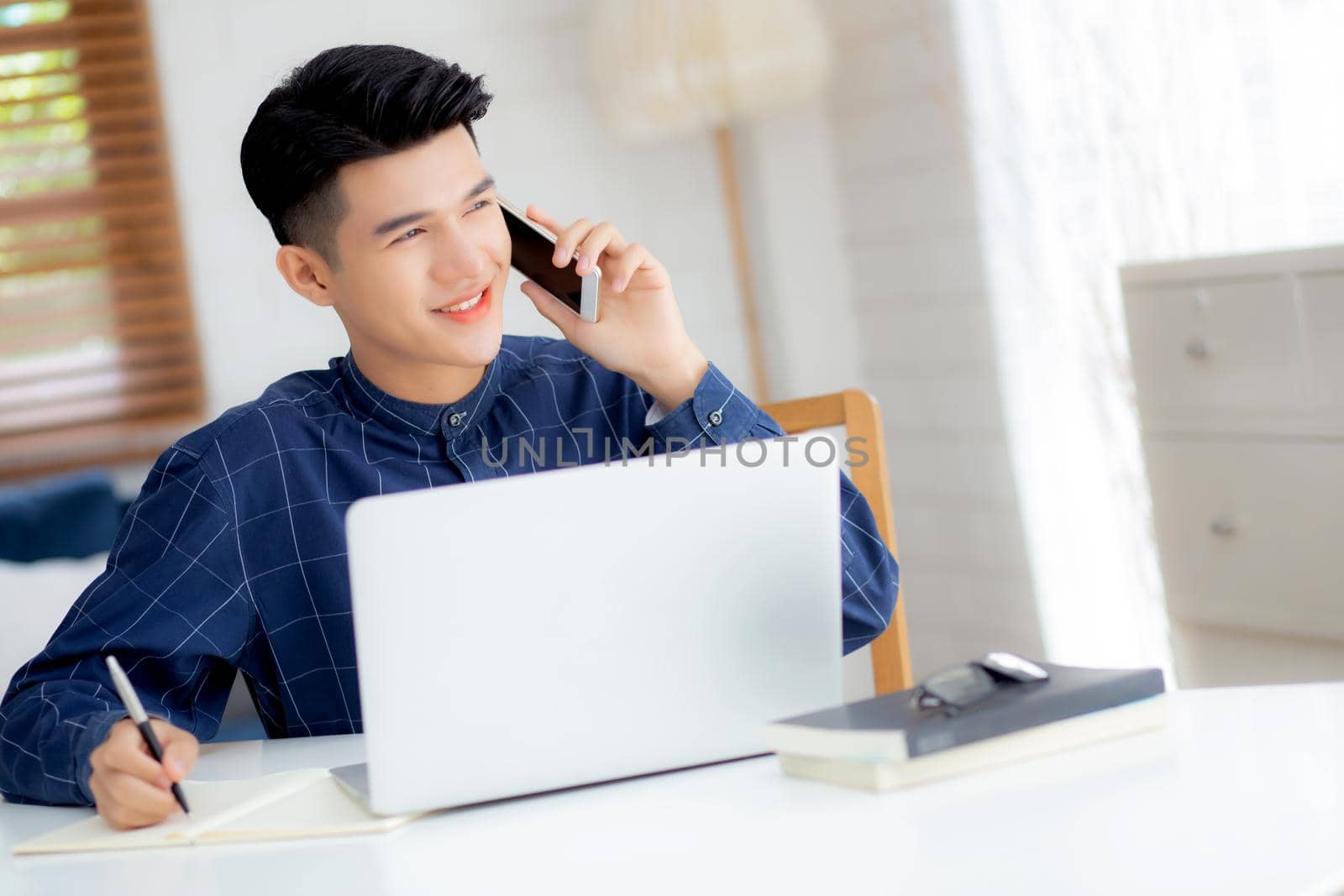 Young asian man talking phone and work from home with laptop computer, freelance male writing on notebook and speak on smartphone, stay home, domestic life, business and communication concept.
