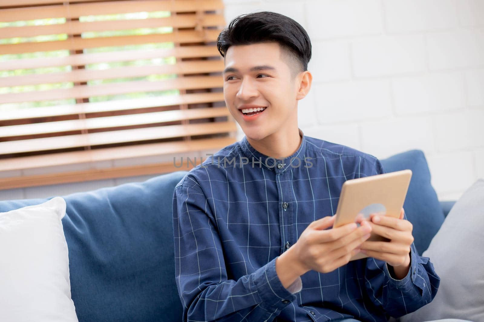 Young attractive asian man resting using browsing tablet computer on sofa at home, happy male sitting on couch relax reading digital gadget at house, communication and lifestyle concept.