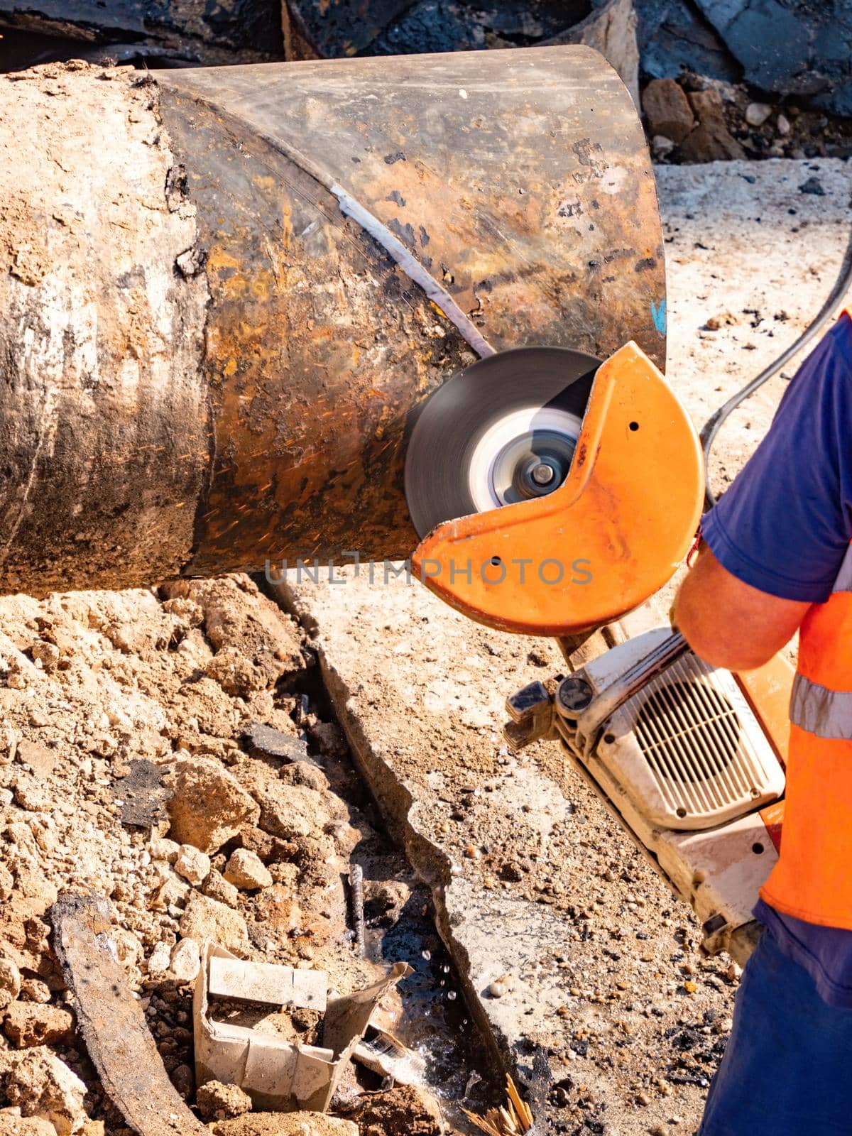 Worker in protective wearing cutting large metal pipe with industrial grinder in  trench