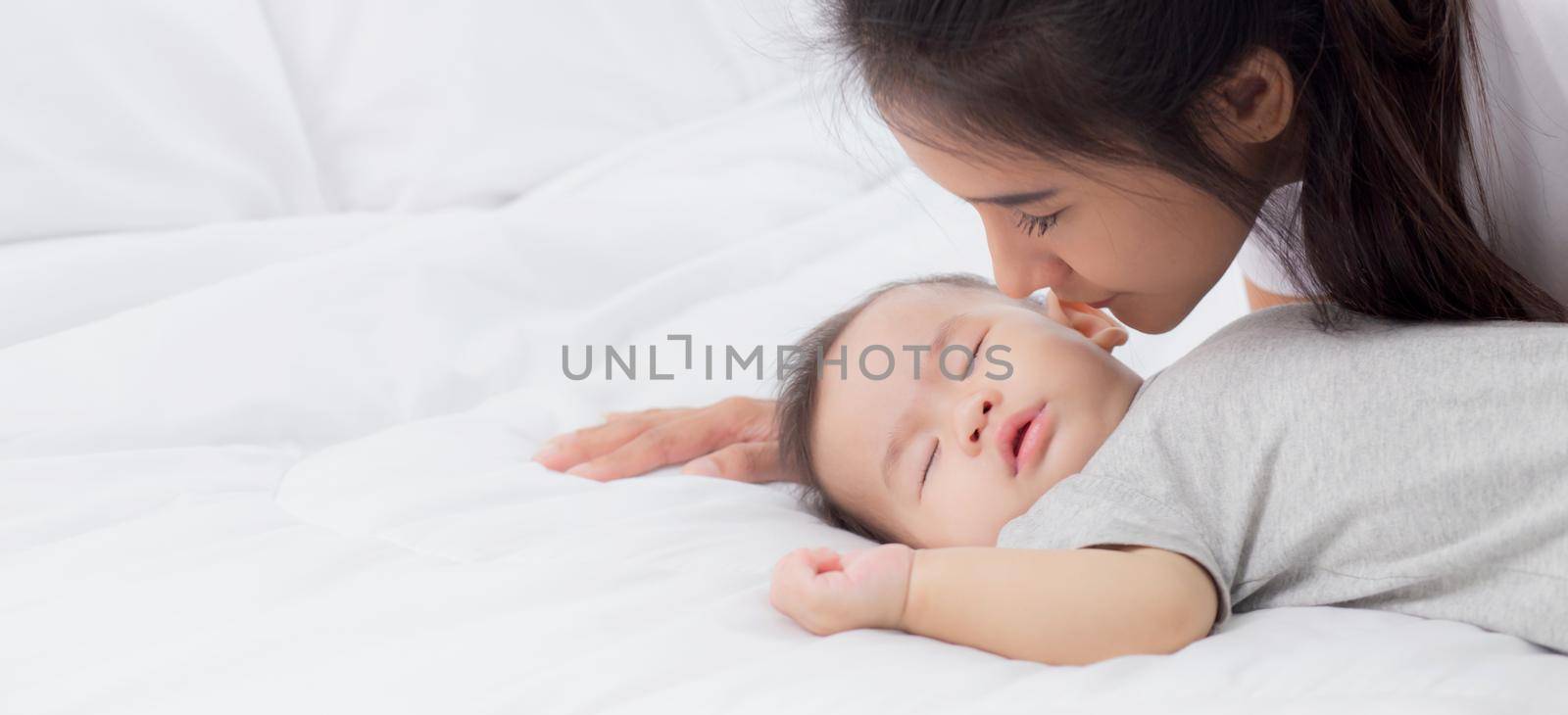 Young asian mother kiss cheek of little baby girl with tender on bed in the bedroom, mom love newborn and care, mother with expression with child together, parent and daughter, family concept. by nnudoo