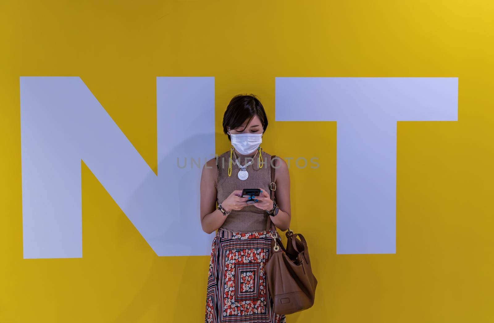 Close-up shot of young asia woman wearing face mask and using a smart phone typing text message while standing in front of yellow wall background during N and T letters. Wears face mask for the coronavirus pandemic. Selective focus.