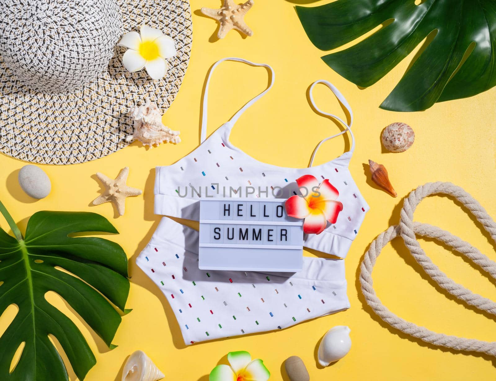 Summer and vacation concept. Words Hello Summer on the lightbox with swimming suit, tropical leaves and seashells flat lay on orange background