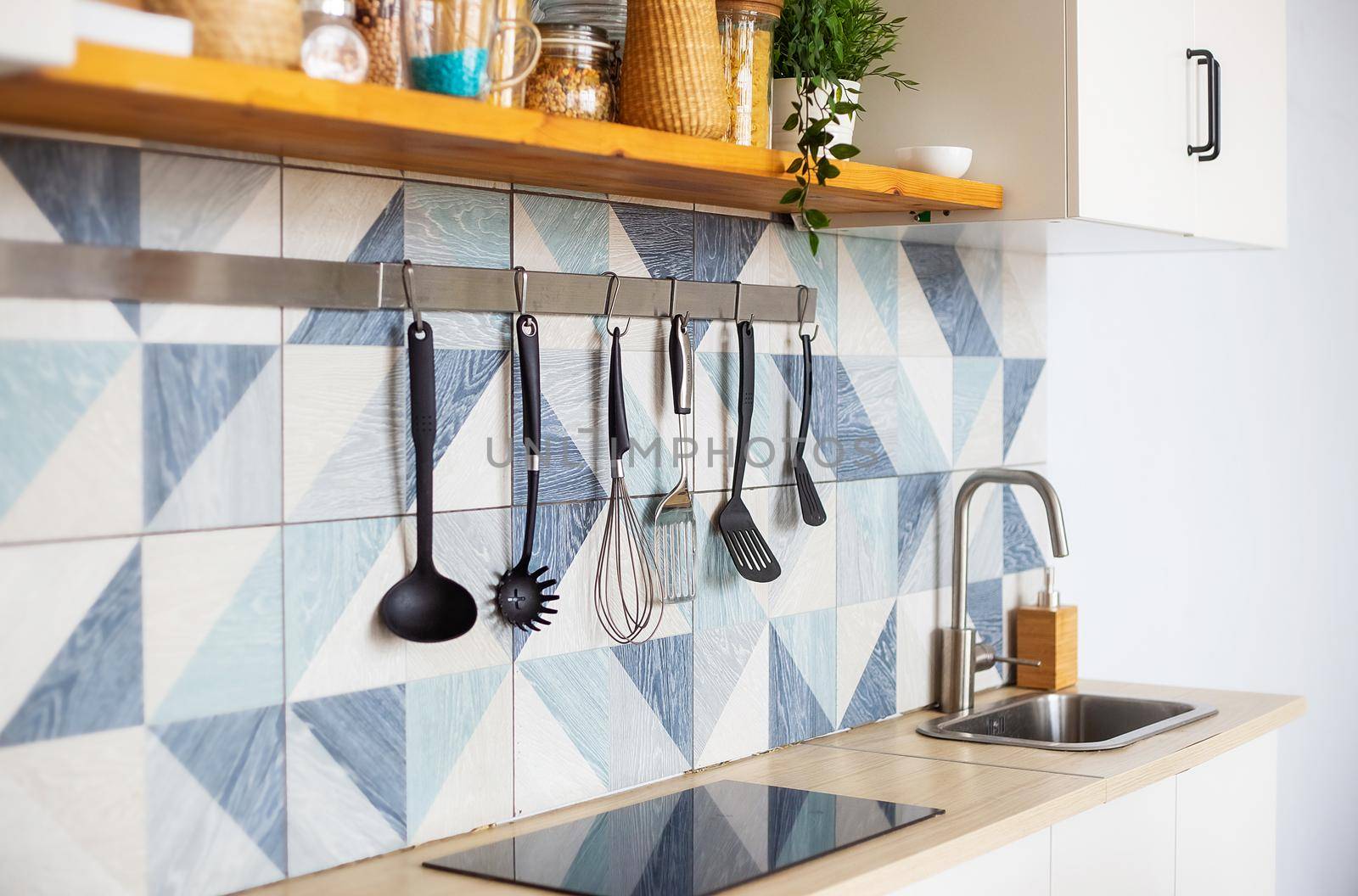 Kitchen spatulas handing on railing on a tiled wall with a blue geometric pattern in the modern designed kitchen room in a Scandinavian stayle. Kitchenware object and interior photo.