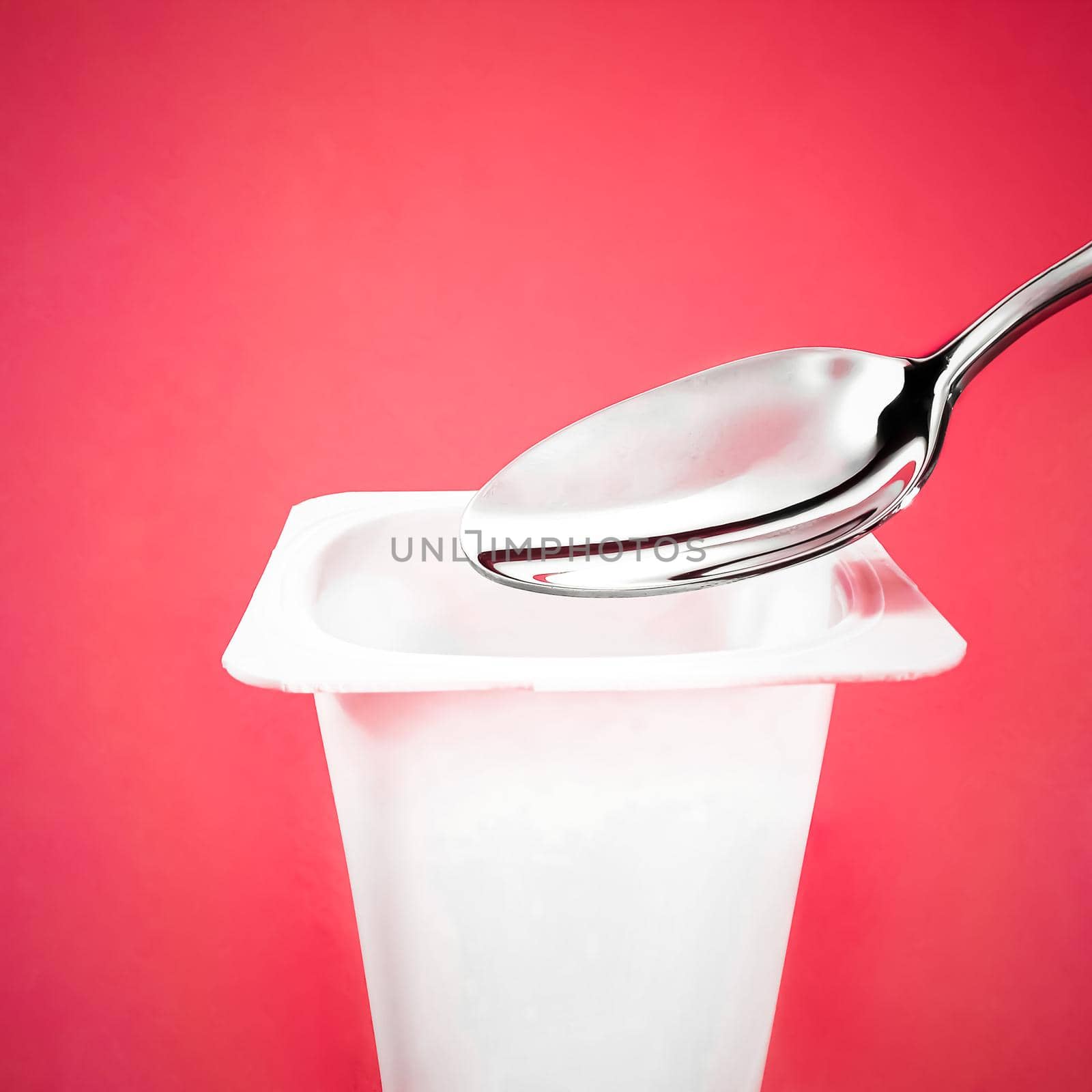 Yogurt cup and silver spoon on red background, white plastic container with yoghurt cream, fresh dairy product for healthy diet and nutrition balance.