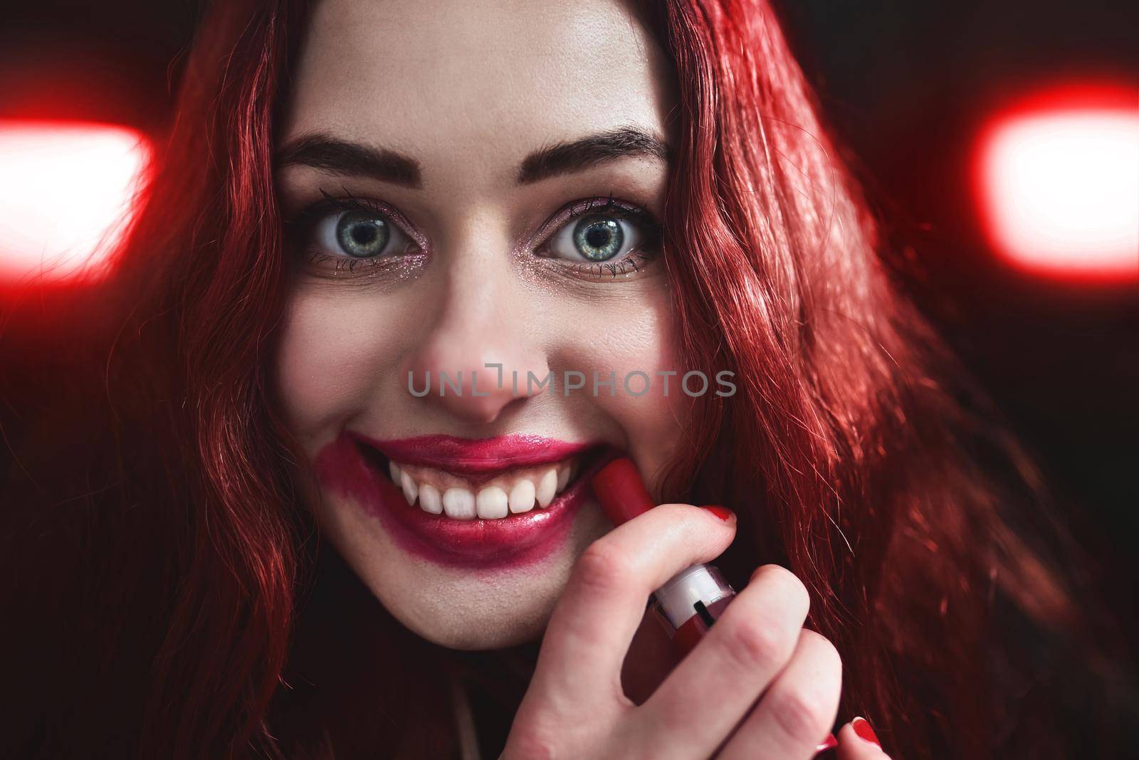 Portrait of crazy-looking teen girl with red hair she is smearing red lipstick on her face, horror concept. halloween time by Nickstock