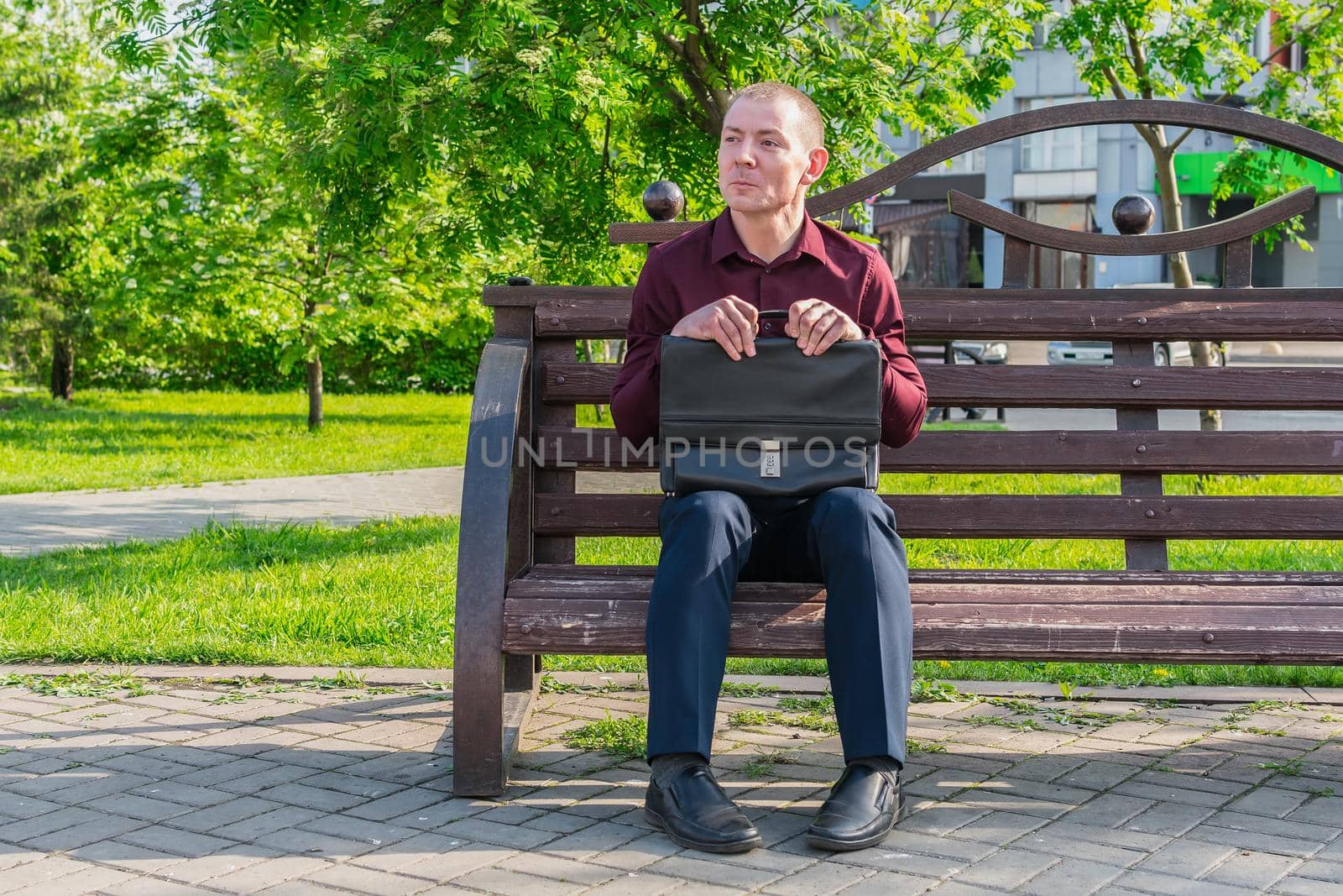 An office worker in a business suit with a suitcase and documents sits with a stupid, confused view on a bench in a city park and expects a meeting