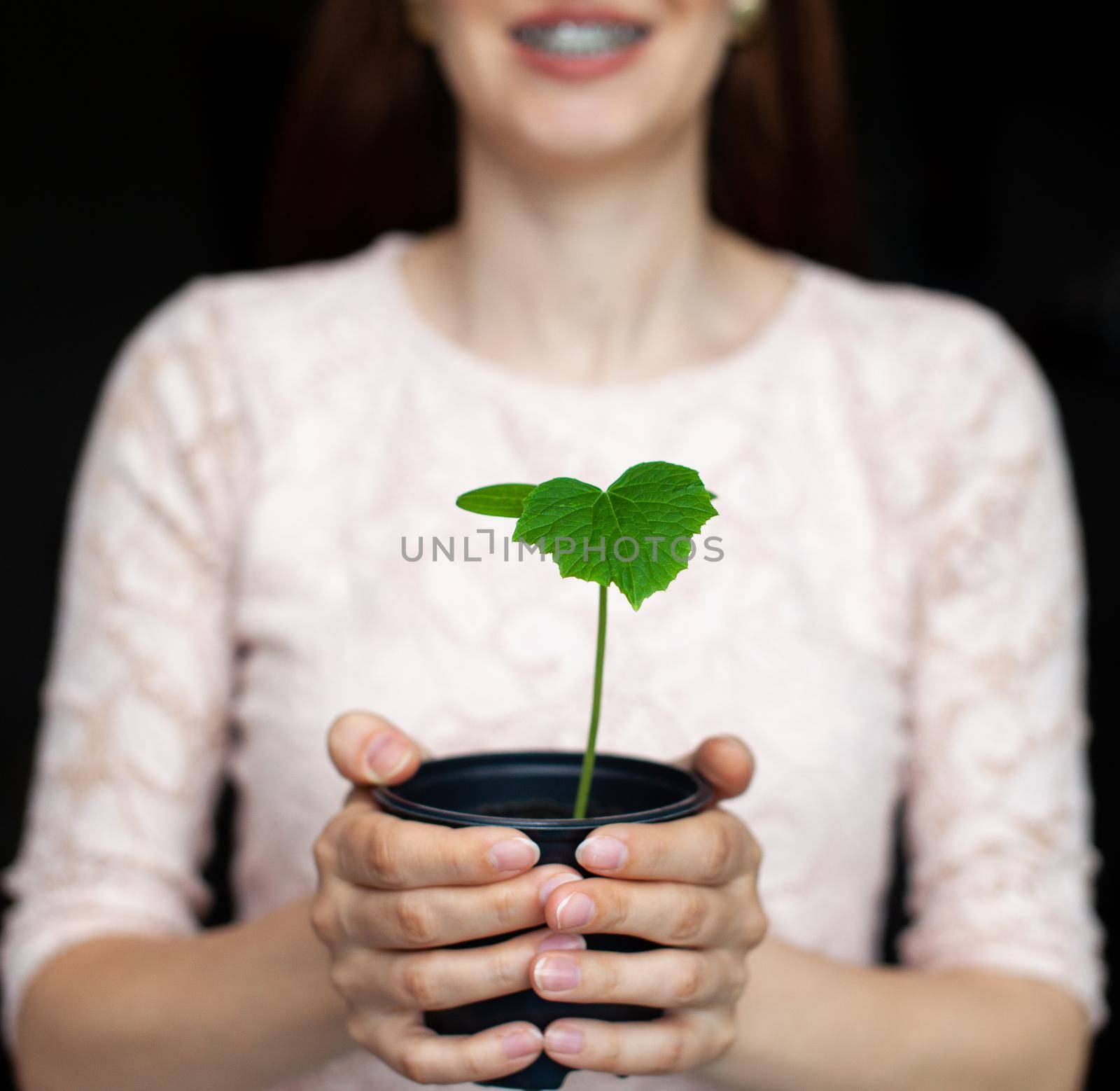 The girl is holding a black pot with a green plant on a dark background. Seedlings of cucumbers in a pot, ready for planting in the ground. Environmental protection. Respect for nature