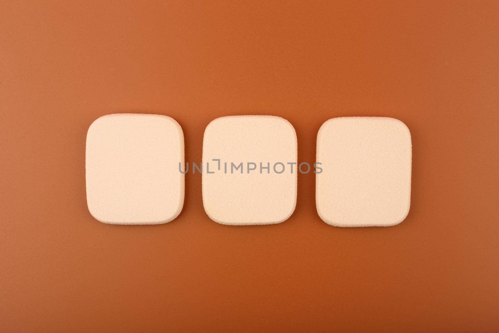 Three beige square shaped make up sponges on brown background. Concept of make up and visage for dark skin tones