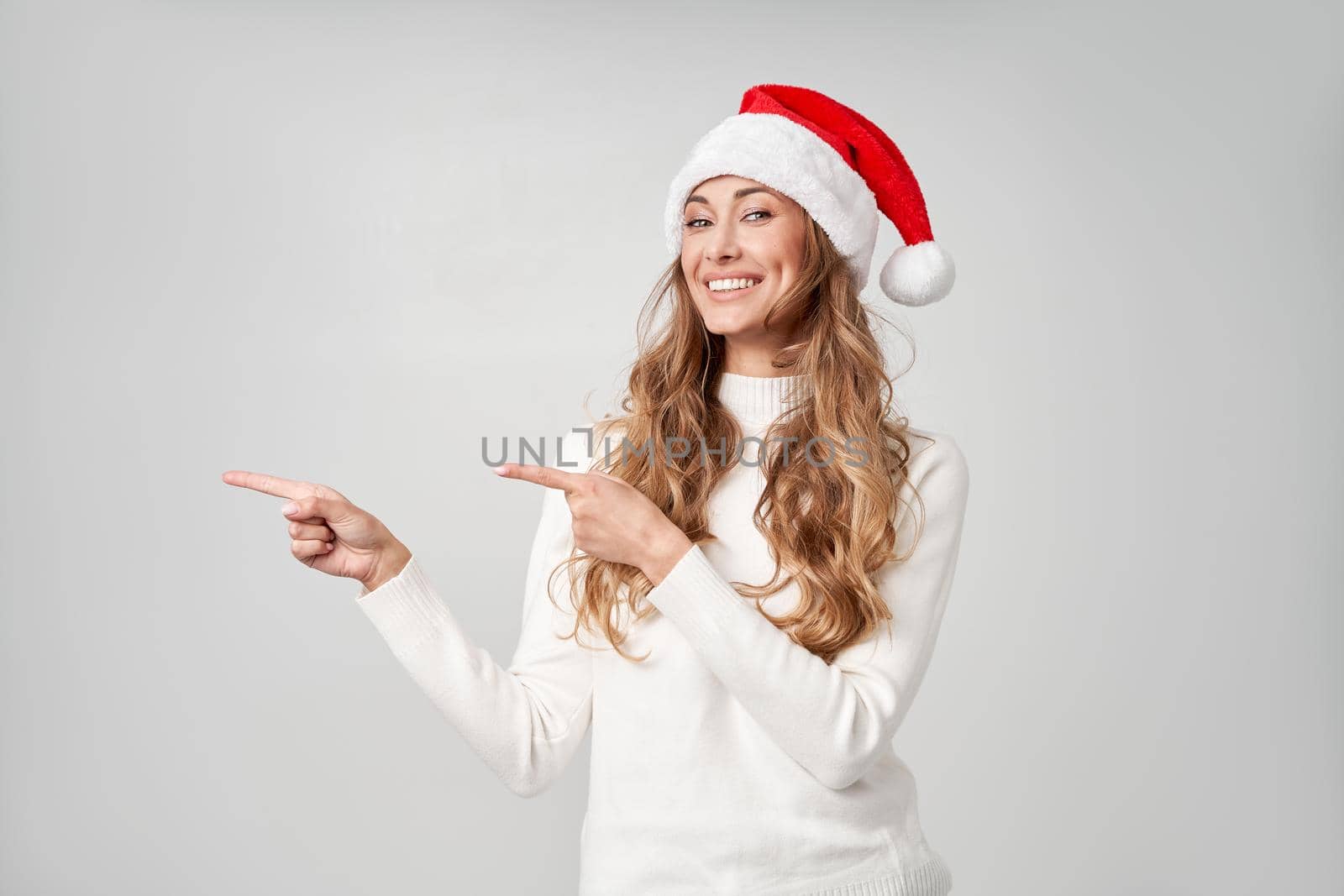 Woman christmas Santa Hat sweater white studio background Beautiful caucasian female curly hair portrait Happy person positive emotion Holiday concept Shows finger on side cope space for advertise