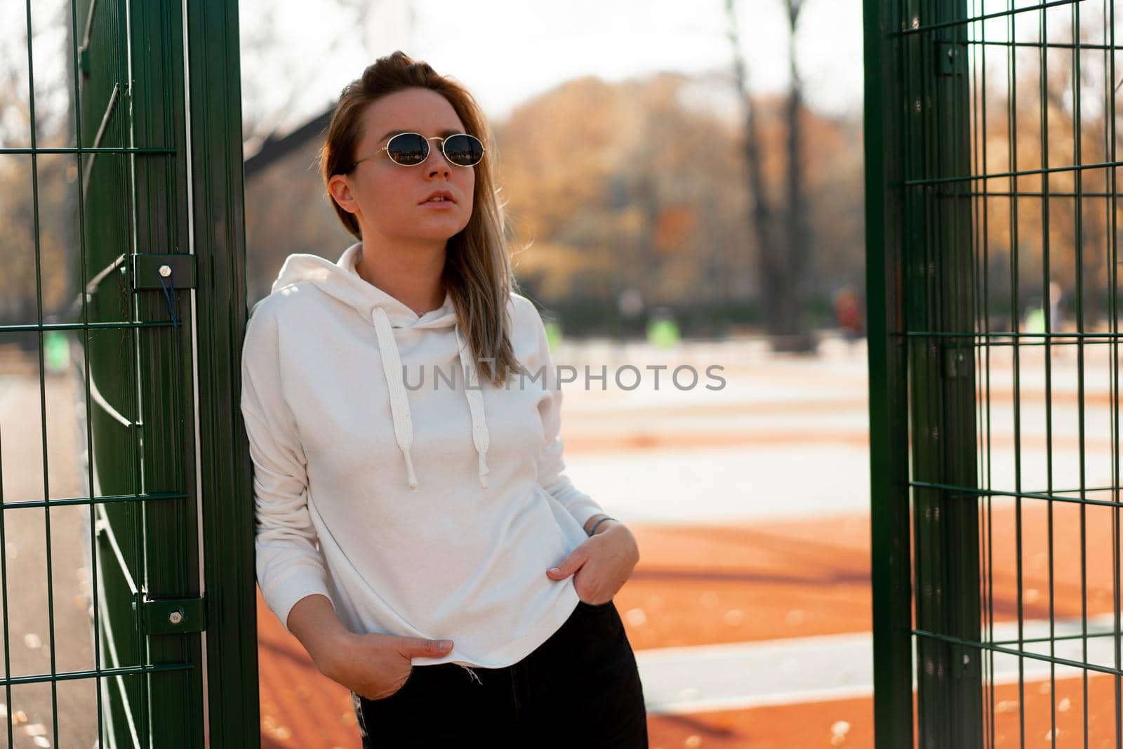 Outdoor close up portrait of young beautiful woman with long hair in sunglasses, dressed in a white hoodie sweater, near the sportsground. youth culture summer pastime