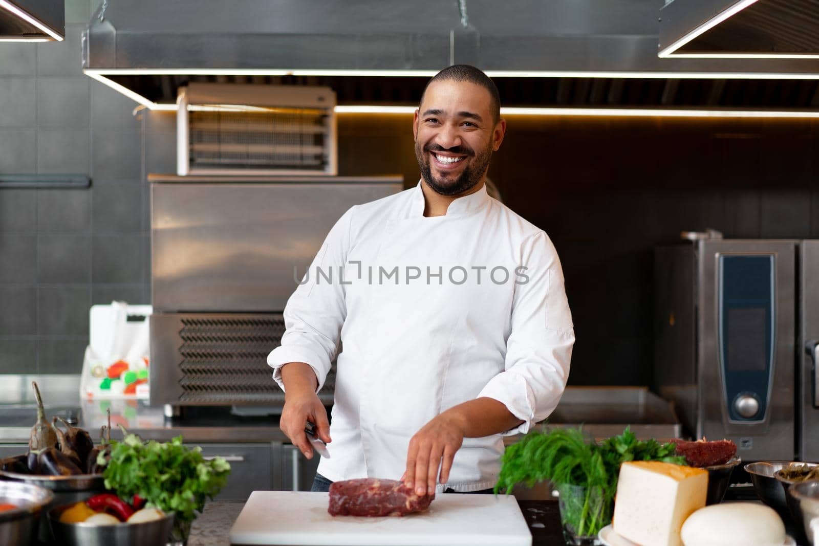 Handsome young African chef standing in professional kitchen in restaurant preparing a meal of meat and cheese vegetables. by andreonegin