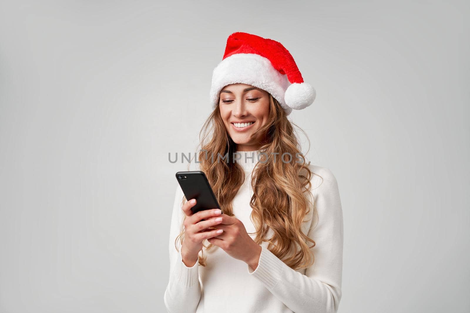 Woman christmas Santa Hat white sweater white studio background with smartphone in hand Beautiful caucasian female curly hair portrait. Happy person positive emotion Holiday concept Teeth smiling