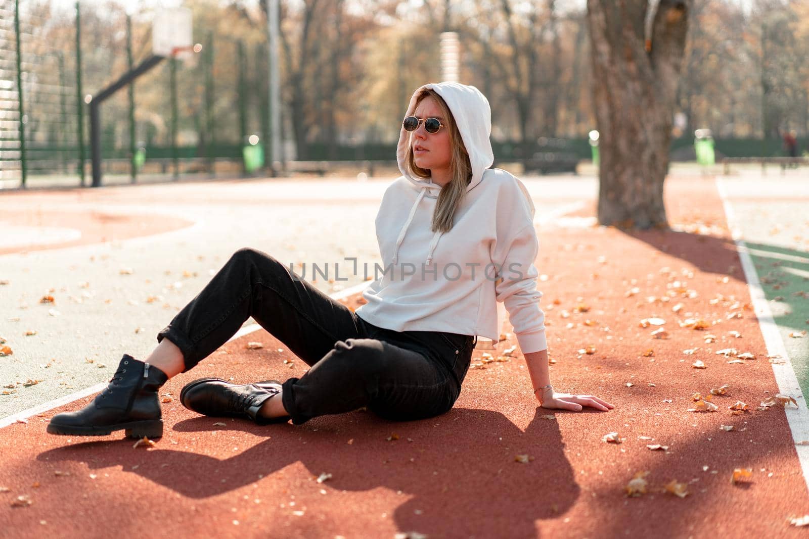Outdoor portrait of young beautiful woman with long in sunglasses and a white hooded sweater sitting on the sportsground track by andreonegin