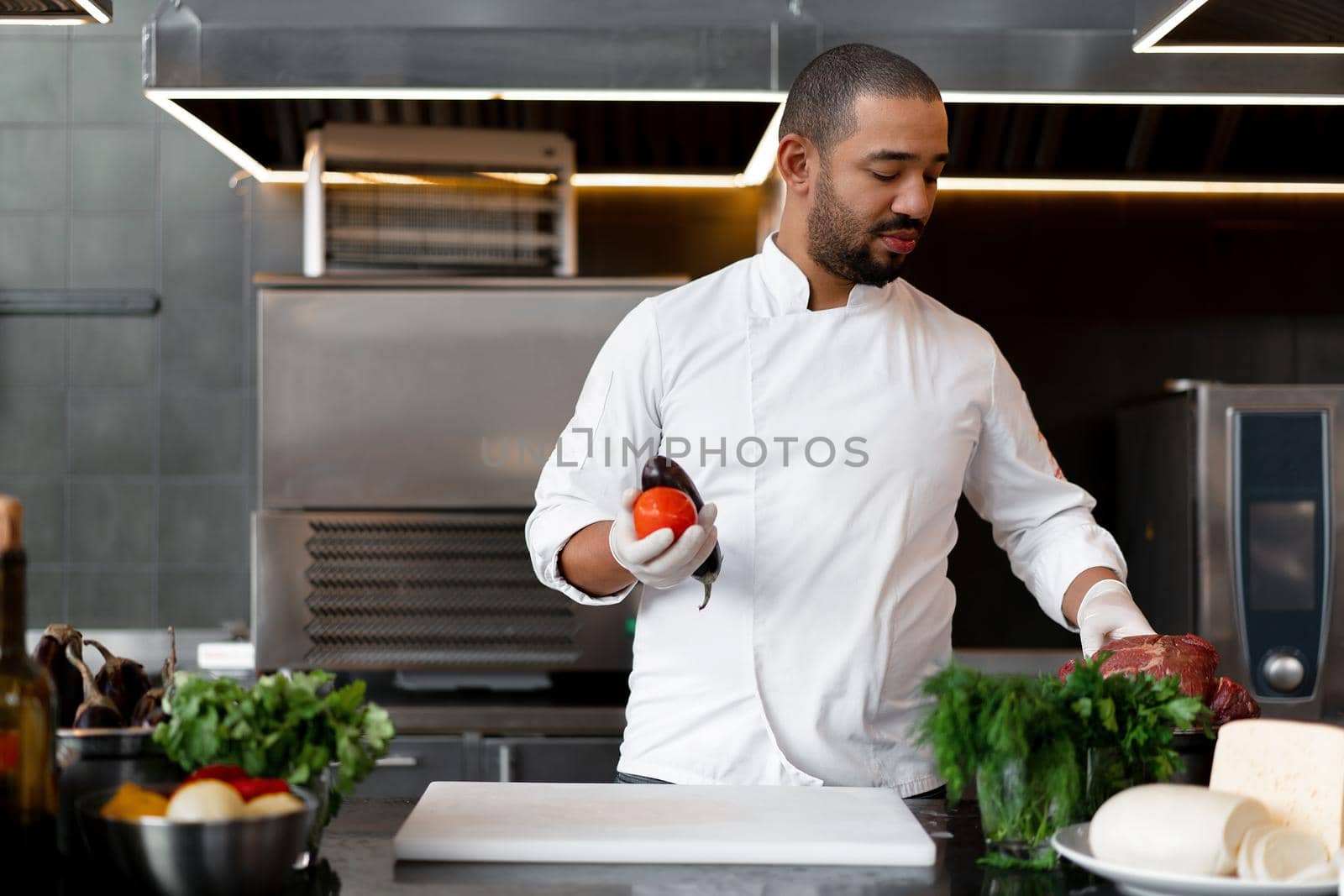 Happy smiling chef prepares meat dish with various vegetables in the kitchen. In one hand the man holds vegetables, in the other a piece of fresh meat by andreonegin