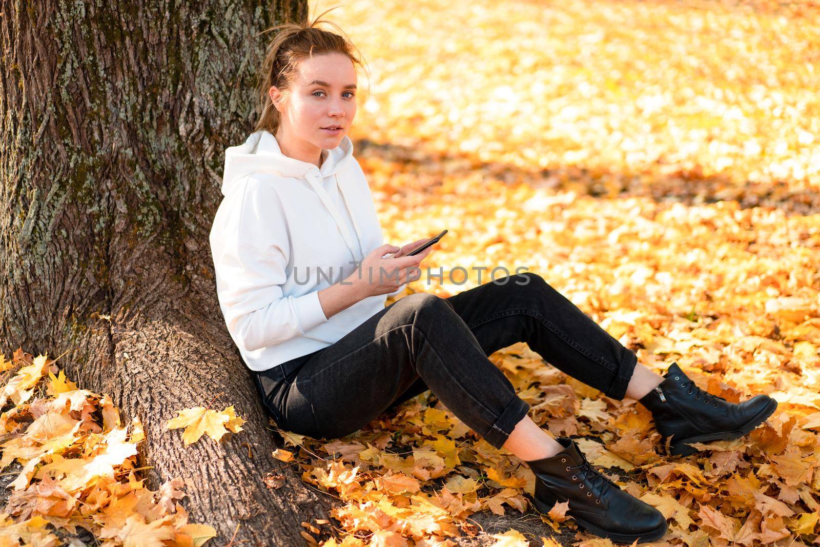 Woman in a white sweater with a hood sits on ground in the park near the tree and holding a cellphone in her hands by andreonegin