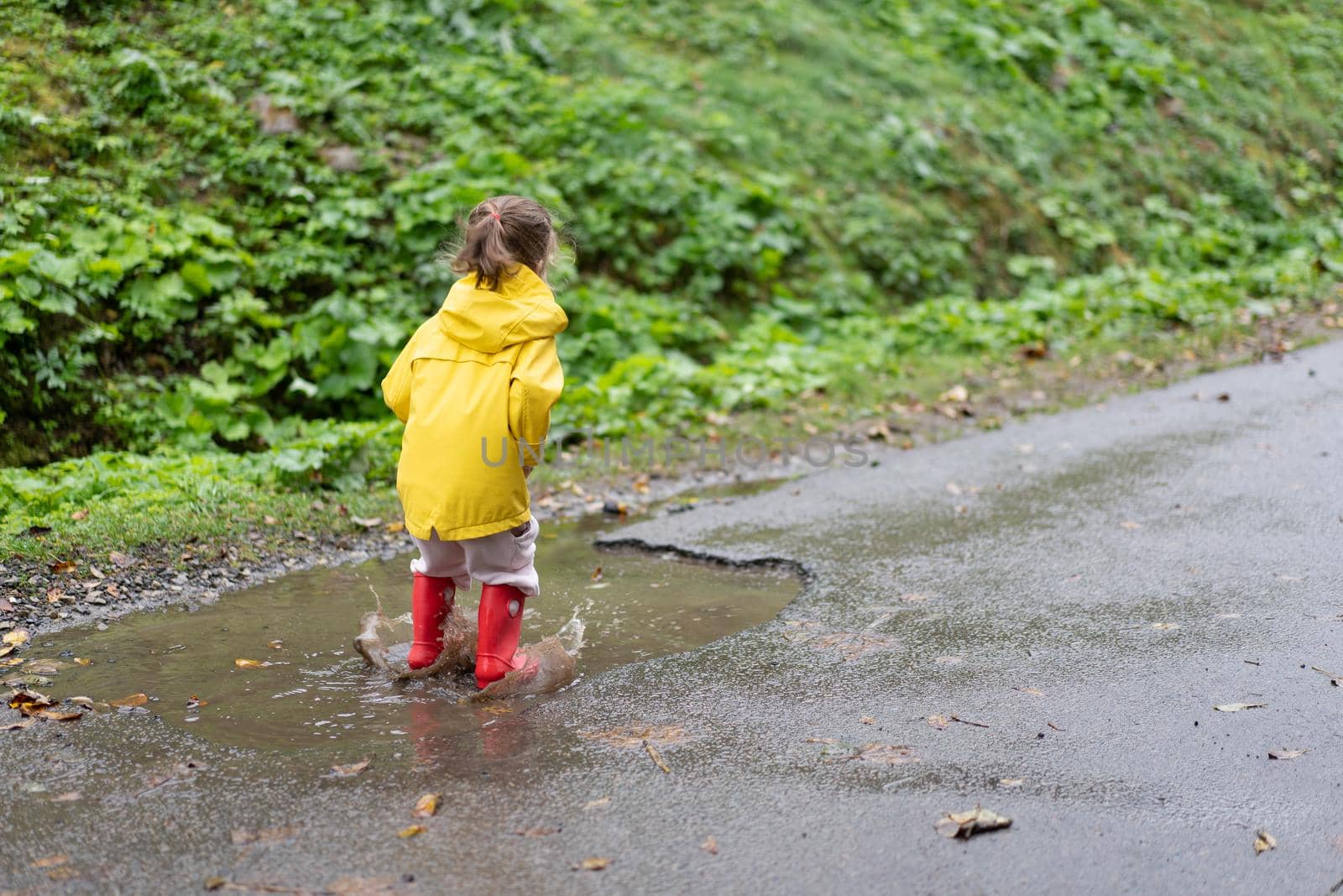 Playful girl wearing yellow raincoat while jumping in puddle during rainfall Happy childhood