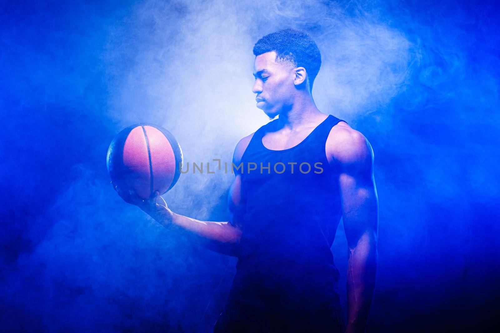 Basketball player holding a ball against blue fog background. African american man looking at basket.