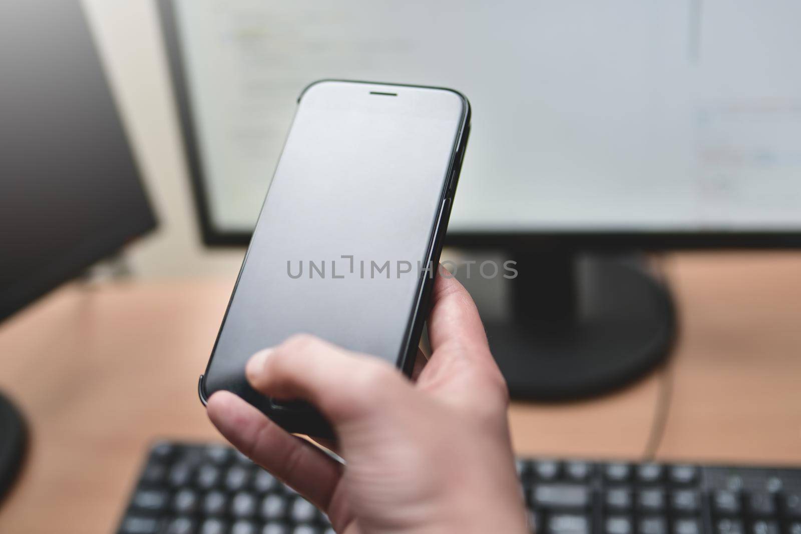 Person holding a smart phone and pointing her finger at the blank screen against a computer keyboard,monitor on wooden table. business concept by Nickstock