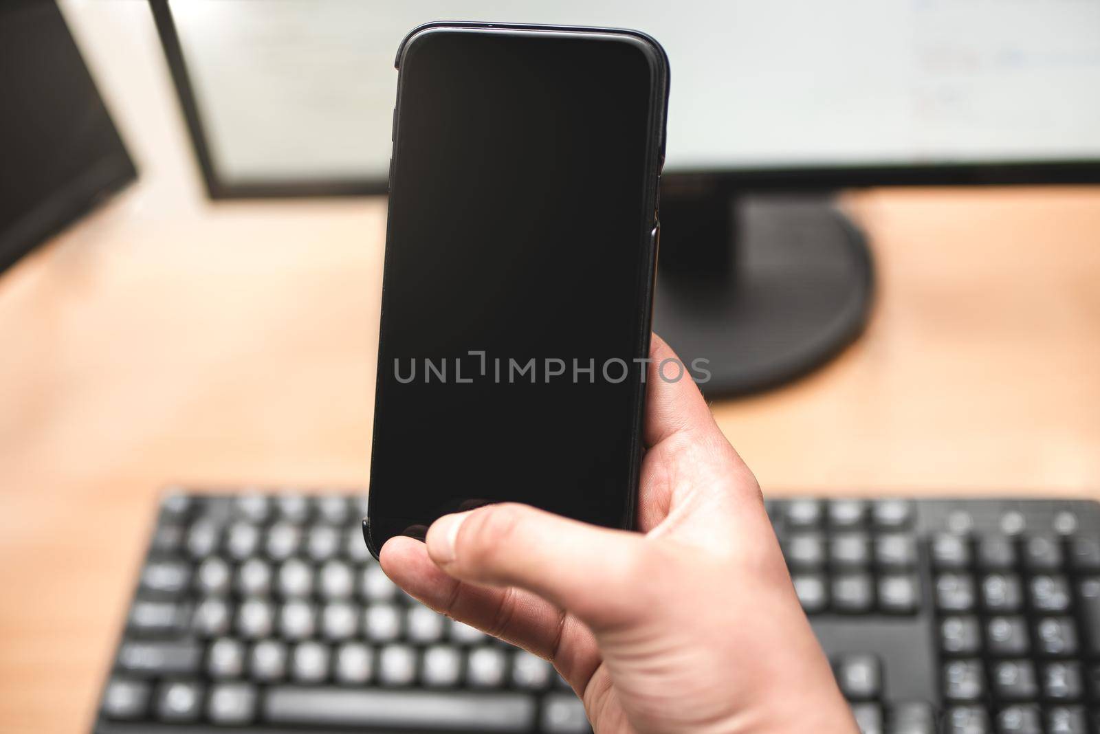 Person holding a smart phone and pointing her finger at the blank screen against a computer keyboard,monitor on wooden table. business concept by Nickstock