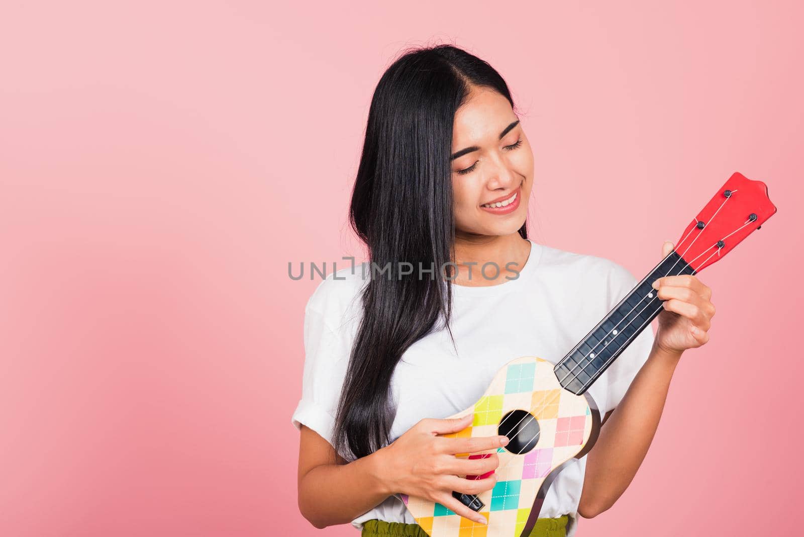 Portrait of happy Asian beautiful young woman teen confident smiling face hold acoustic Ukulele guitar, female playing Hawaiian small guitar, studio shot isolated on pink background, with copy space