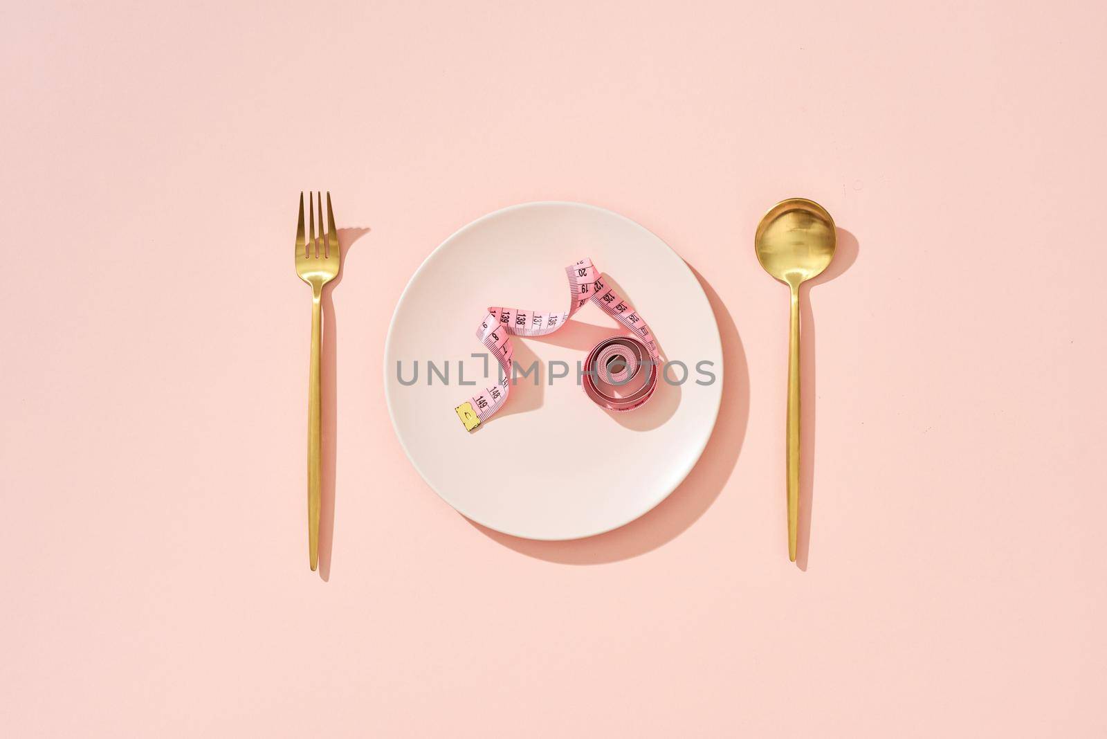 Roll of measuring tape on a pink dinner plate, table knife and fork on a pink background. by makidotvn