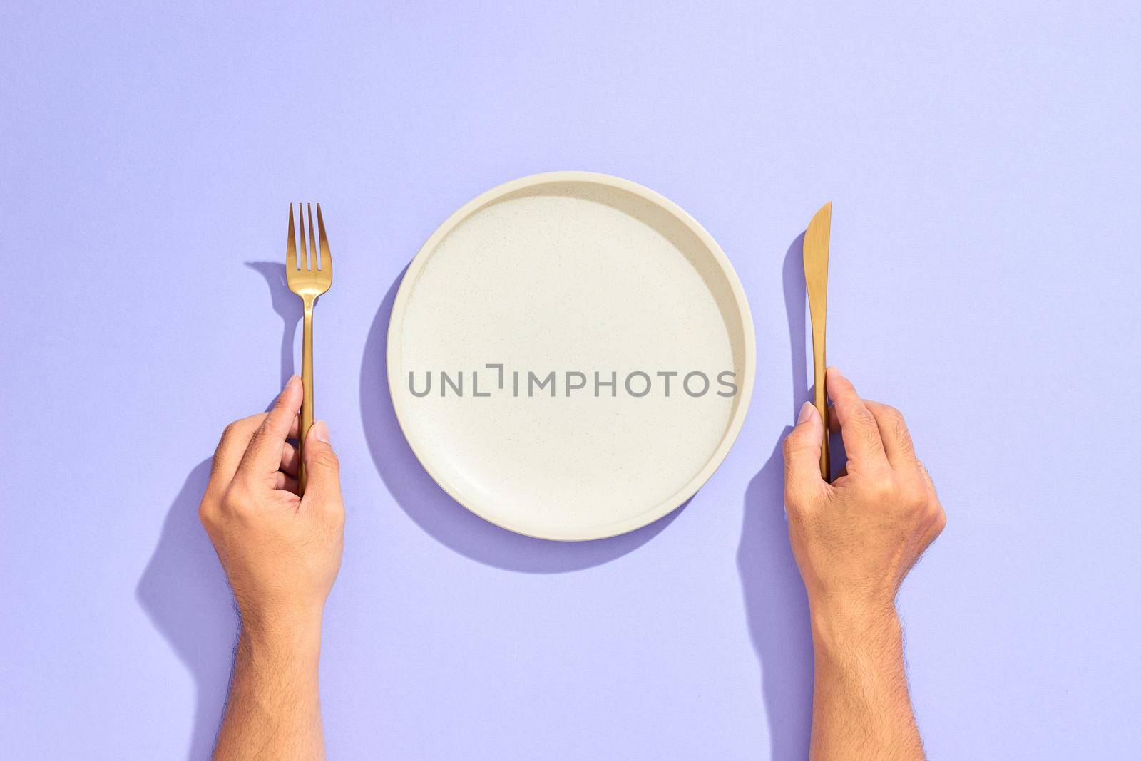Dinner place setting. White empty plate and hand holding gold fork and knife on lilac background by makidotvn