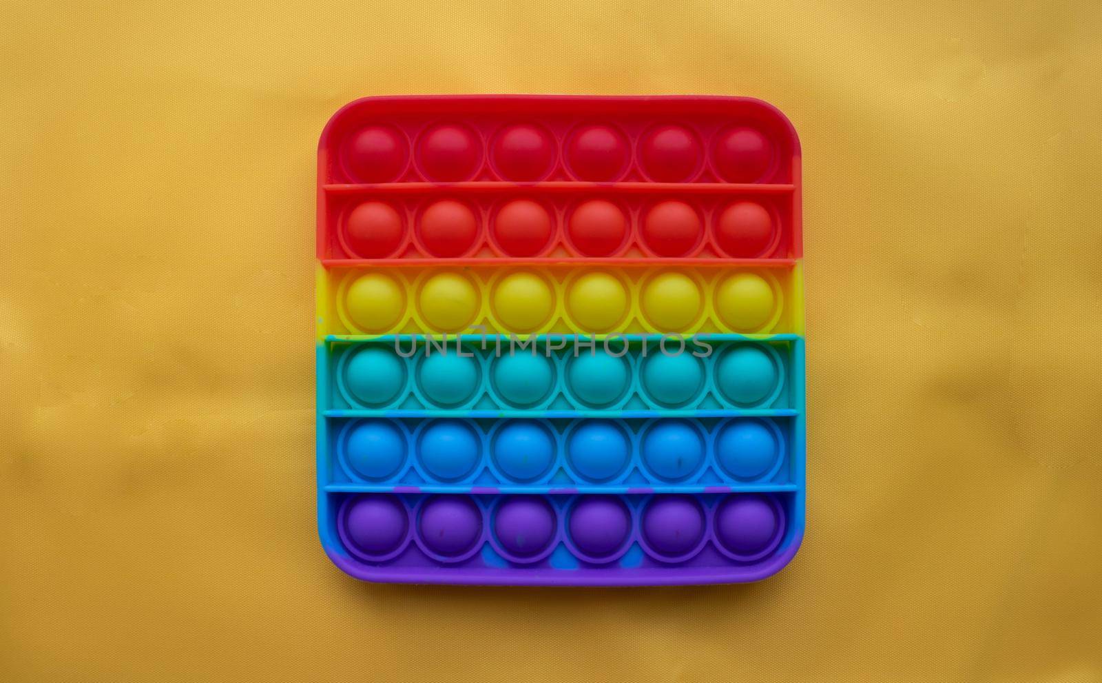 A rainbow toy lies against a background of yellow fabric. Blue and Orange Funny Interactive Anti-Stress Silicone Toys. Pop it.