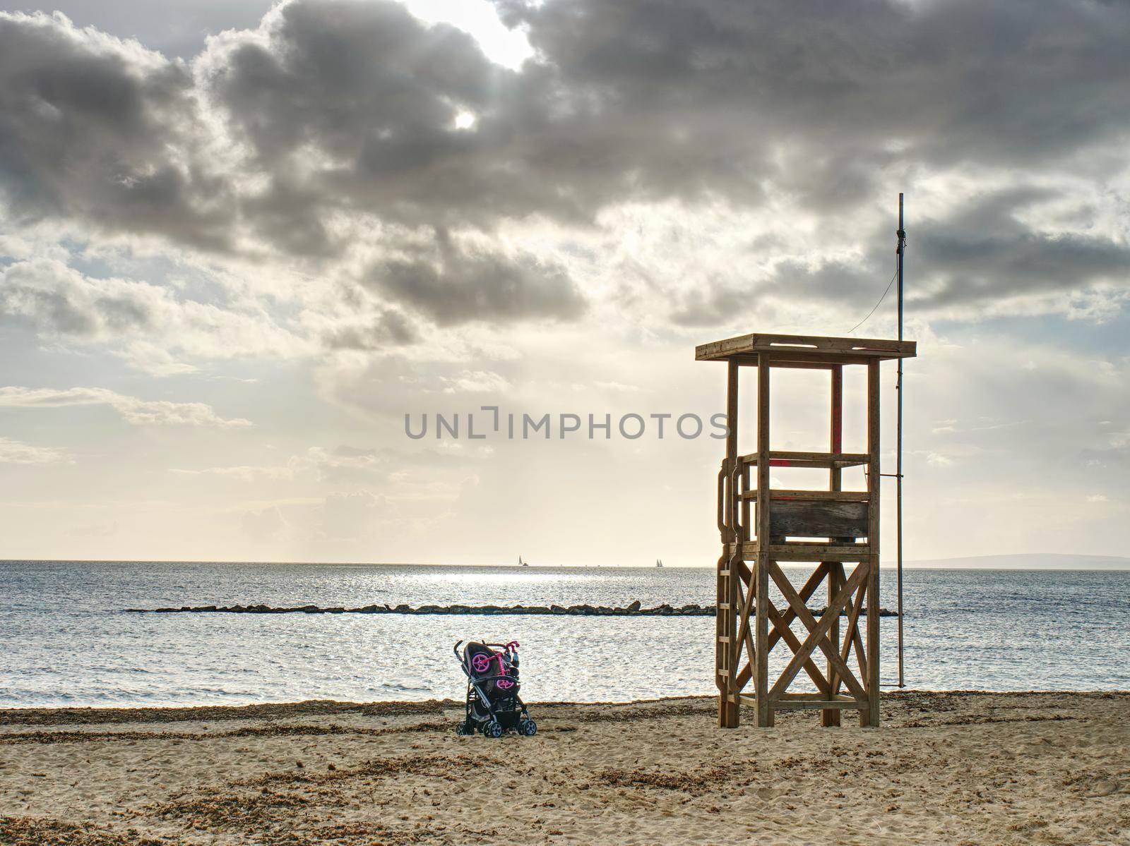 Baby carriage standing under lifeguard wooden tower on empty afternoon sand beach, calm bay level in background.