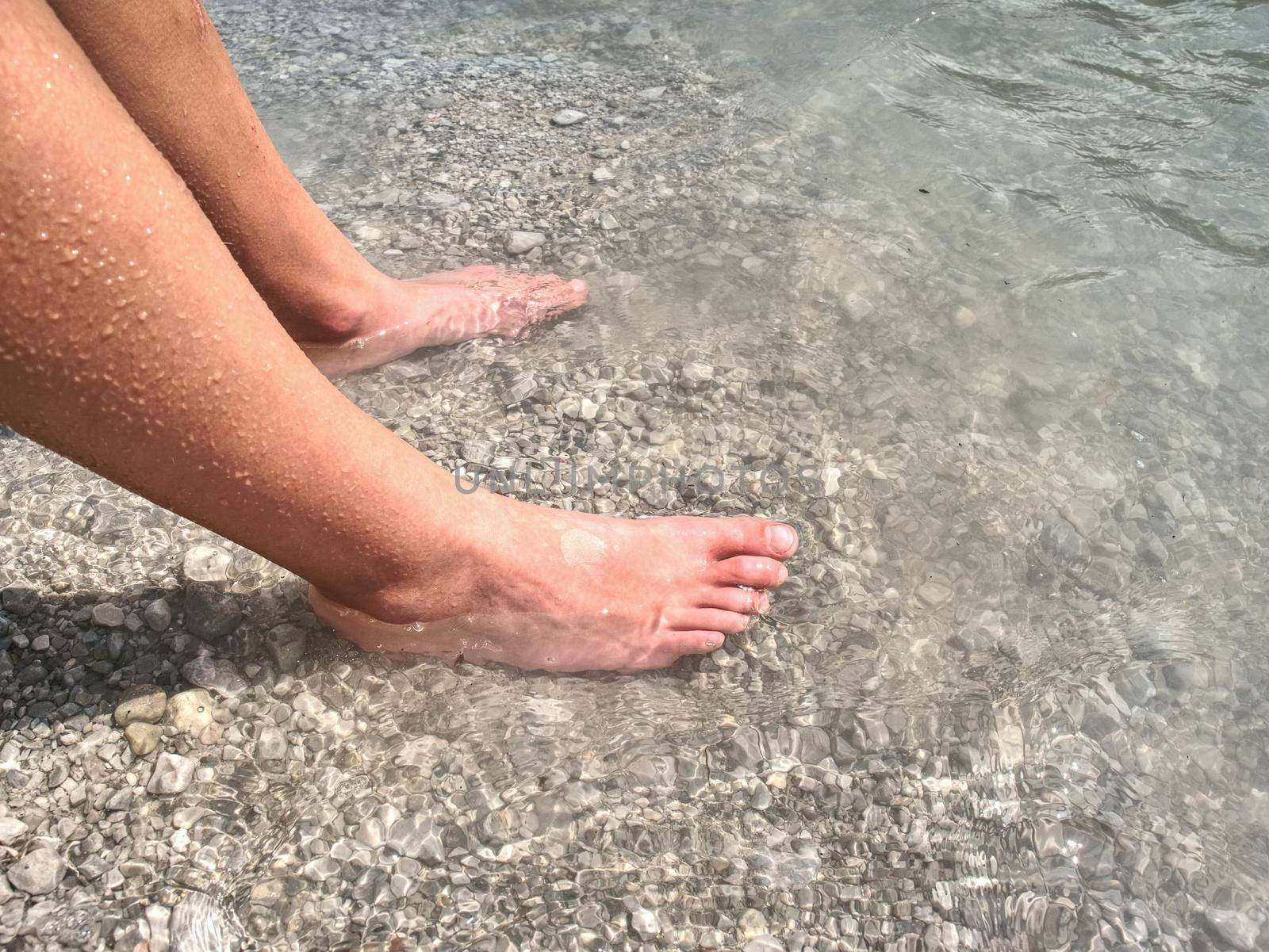 Naked legs along wave of lake water and rough white stony sand on the beach. Carefree and relaxing attitude