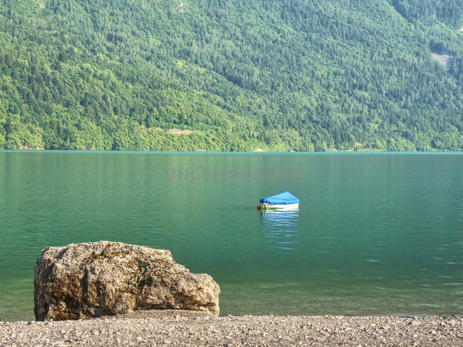 Boat on  green blue water level of Alps lake. White stony shore of a mountain lake. Reflection of forest mountains in water level.