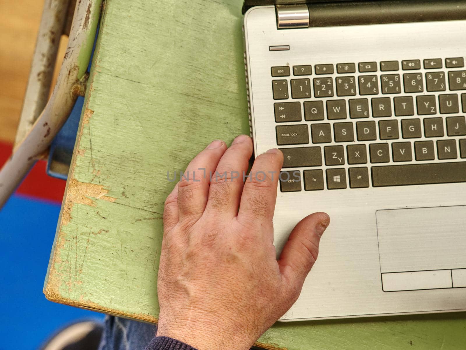 Old male freelancer typing messages via email using laptop computer and mobile phone for distant work while sitting at  worn out desk.