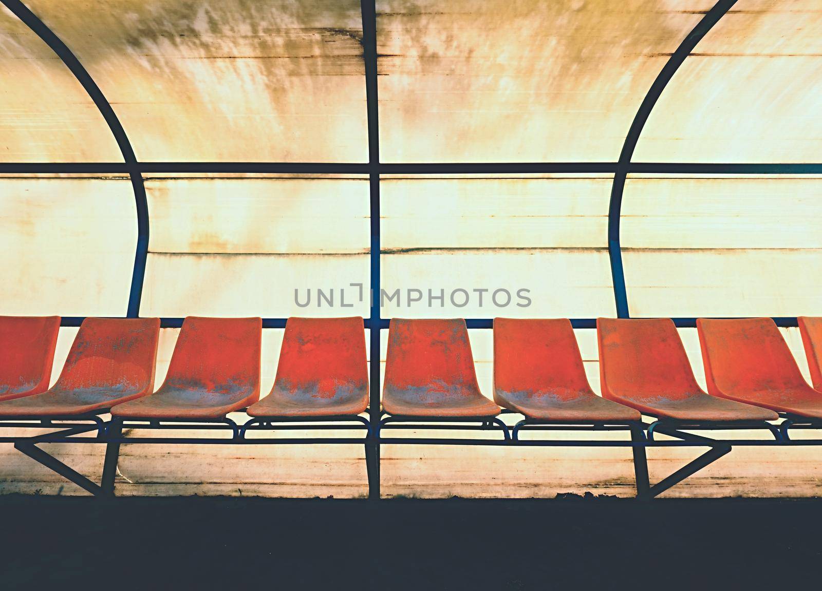 Vintage plastic seats on outdoor stadium players bench, chairs with worn paint below yellow roof.