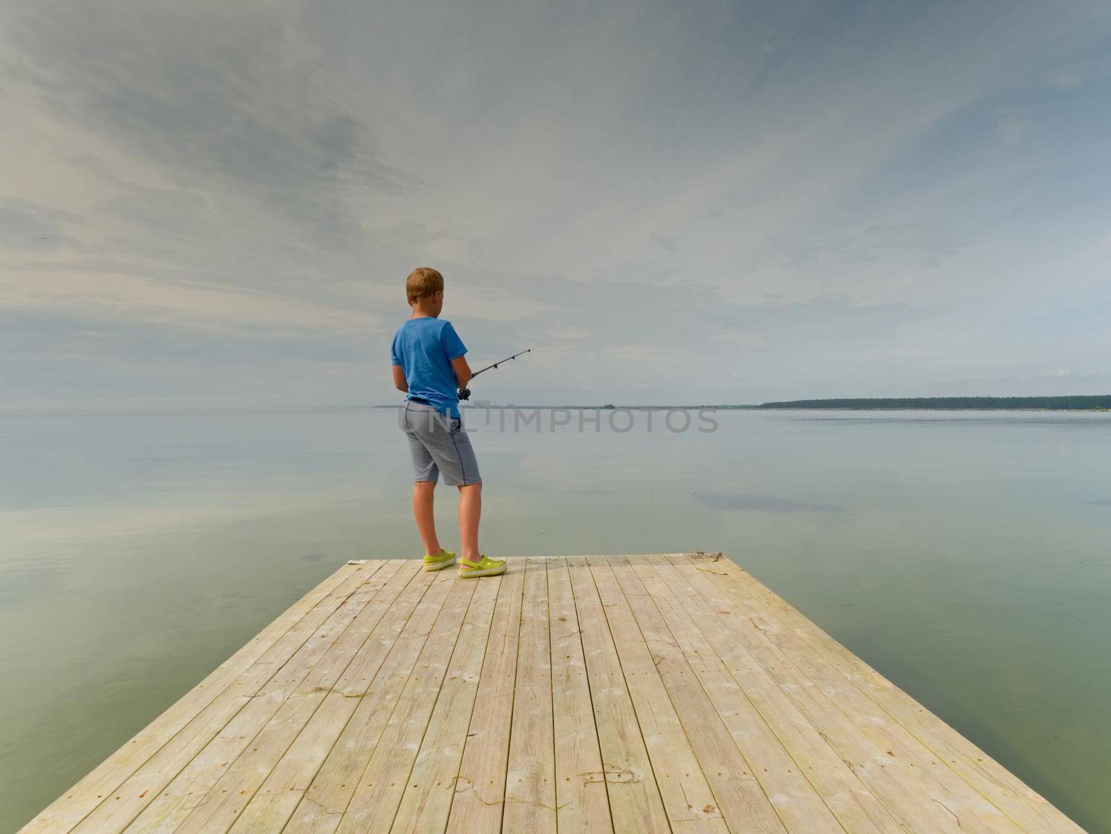 Small blond hair boy is fishing at the end of wooden mole. Smooth water level in bay by rdonar2
