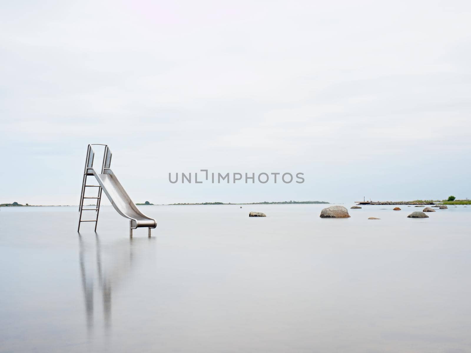 Sunset shore and old steel slide in lake. Chrome ladder tower with  sliding track, big granite stones around. Cold autumn morning with smooth water level and island at horizon.