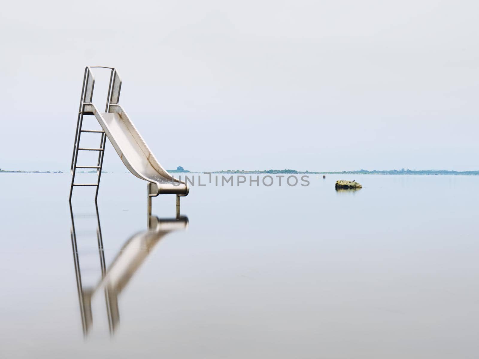 Old steel slide in lake. Chrome ladder tower with  sliding track, big granite stones around. Smooth water level and island at horizon. End of season in  natural swimming resort