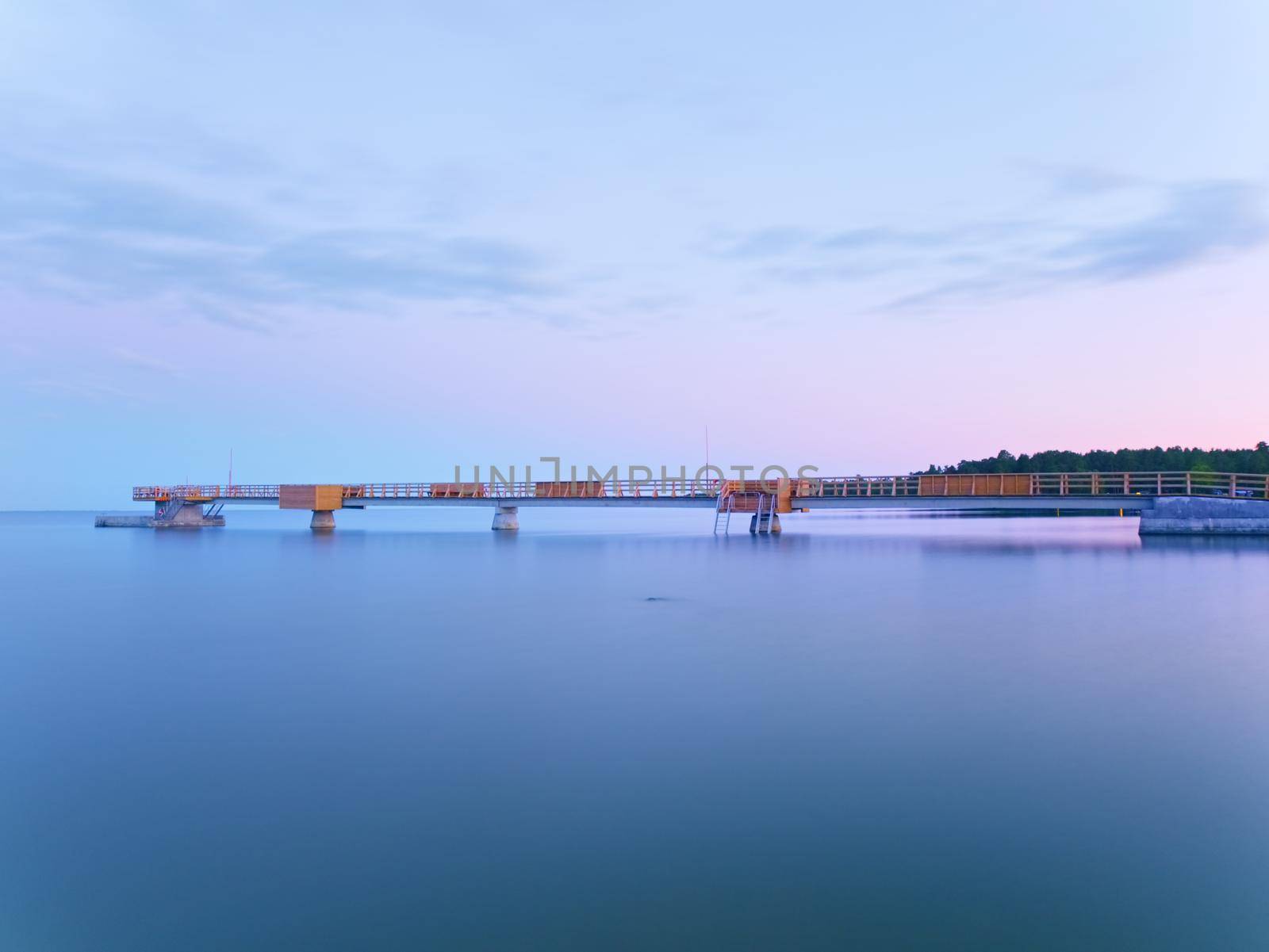 Long wooden sea bridge on coast during morning. Cold weather with first sun rays at horizon, smooth water. New built  pier in summer holiday resort. Empty mole.